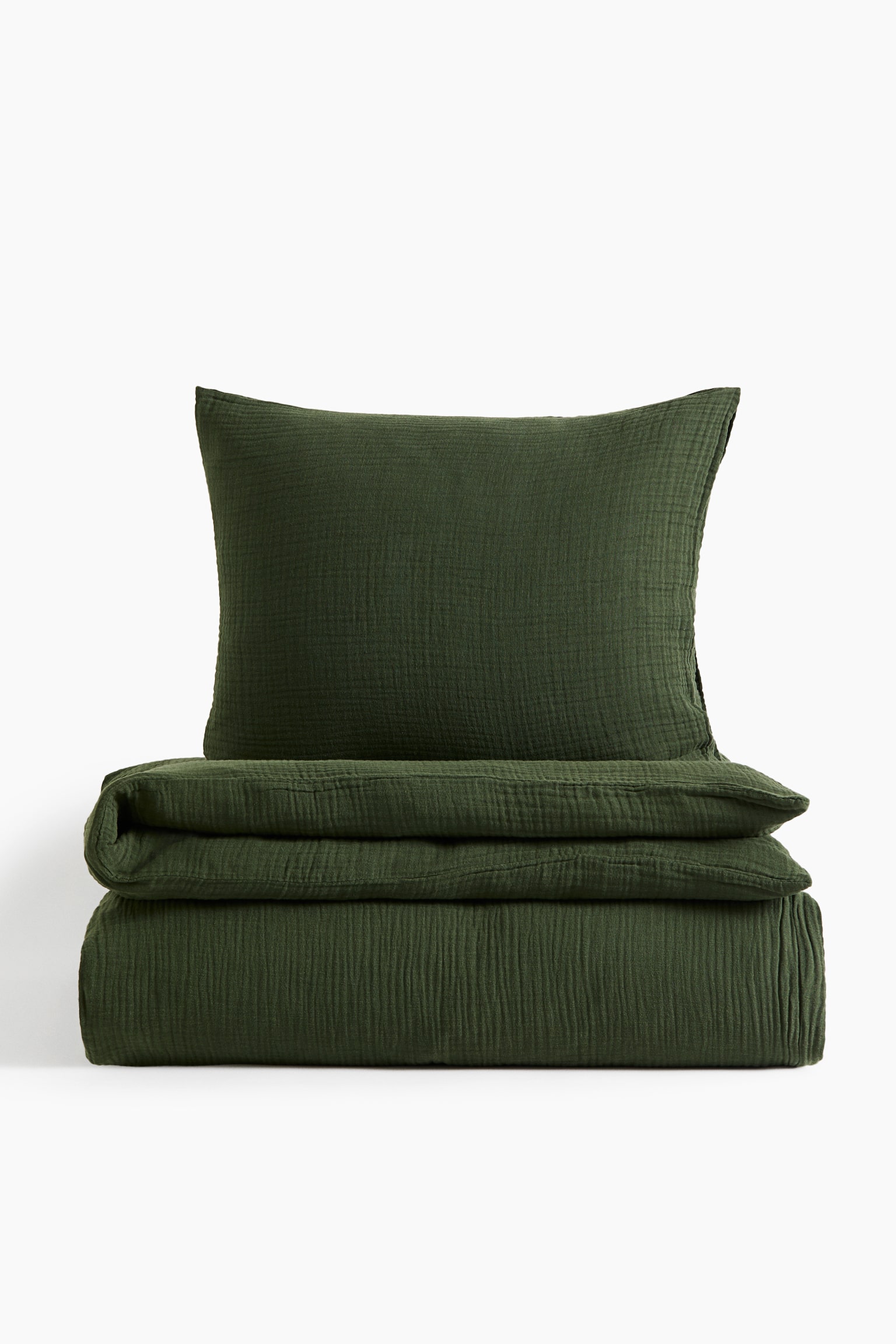A dark green cotton muslin duvet cover set is shown from the front, featuring a neatly folded duvet cover resting at the bottom with a matching pillowcase standing upright behind it. Both pieces display a textured, crinkled appearance.