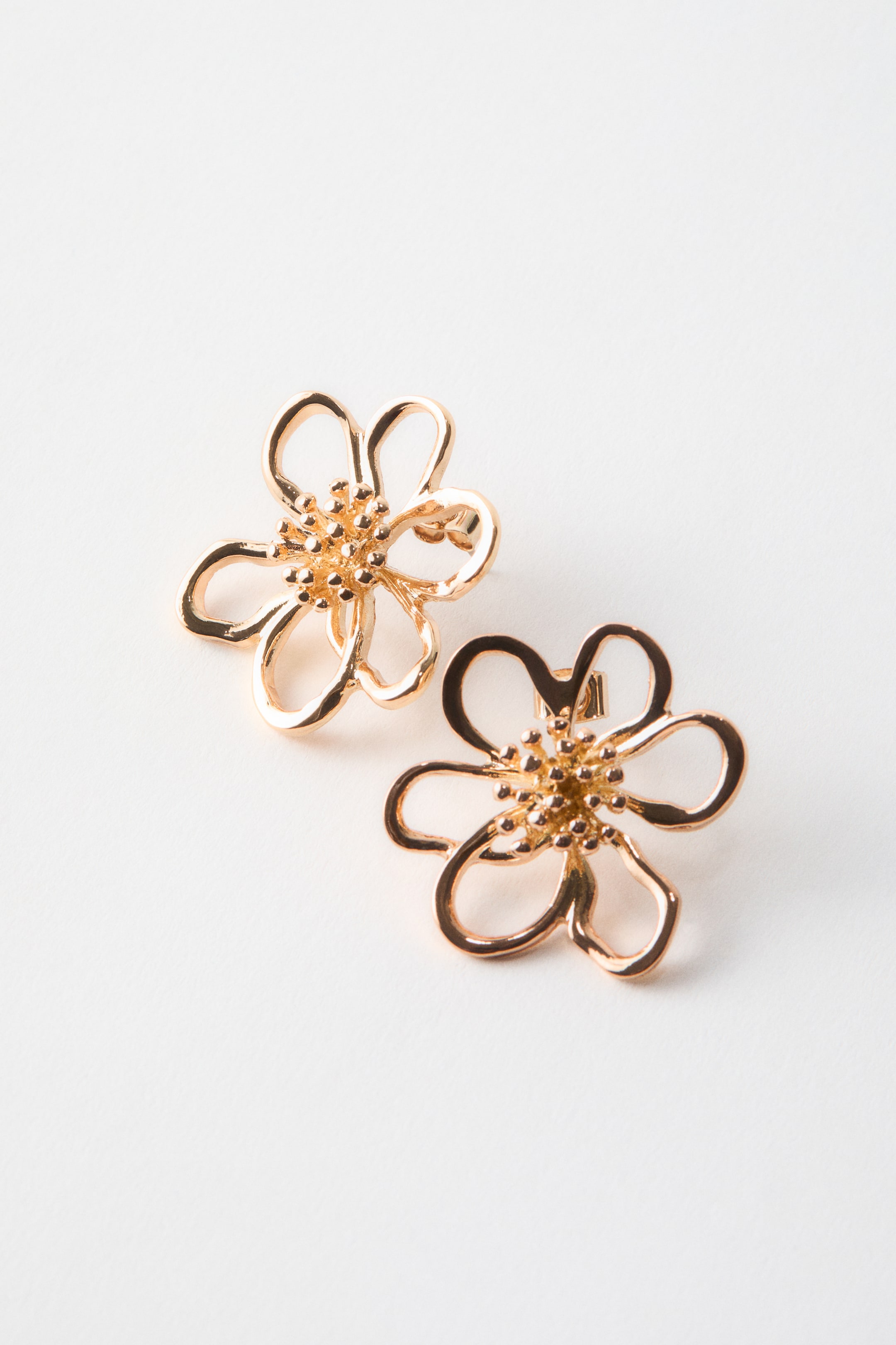View larger image: Two flower-shaped earrings feature a rose gold finish, slightly angled to highlight their three-dimensional structure. Each earring showcases five open, outline petals encircling a textured, beaded center.