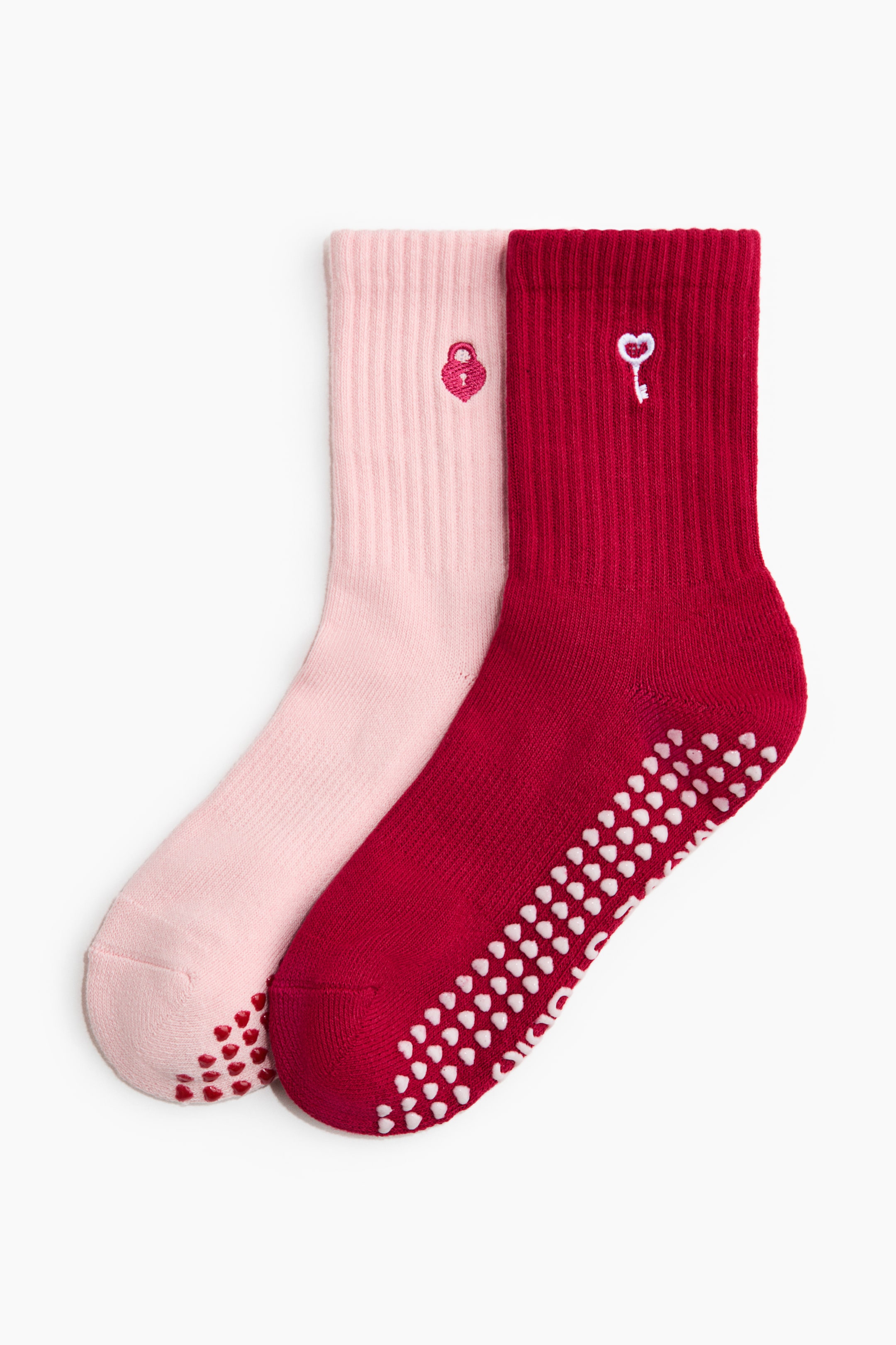 View larger image: Two socks are shown together: a light-pink sock with a red embroidered heart-shaped padlock on its ribbed cuff, and a dark-red sock with a white embroidered key on its ribbed cuff. Both feature heart-shaped non-slip grips on their soles; the grips on the pink sock are red, and those on the red sock are white.
