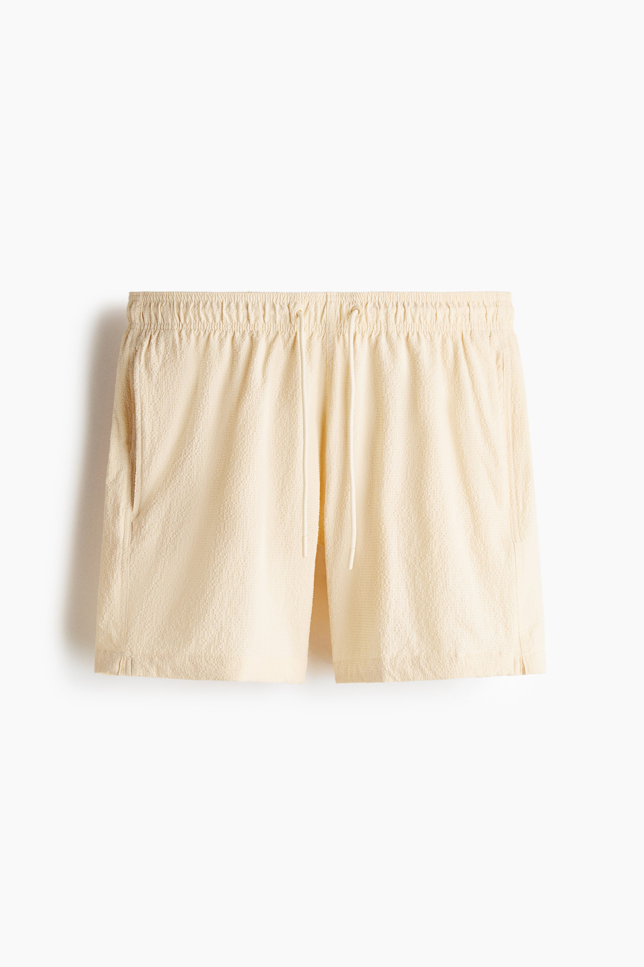 View larger image: Cream textured shorts are shown from the front, with an elastic drawstring waistband, slanted side pockets, and small side slits at the straight hem.