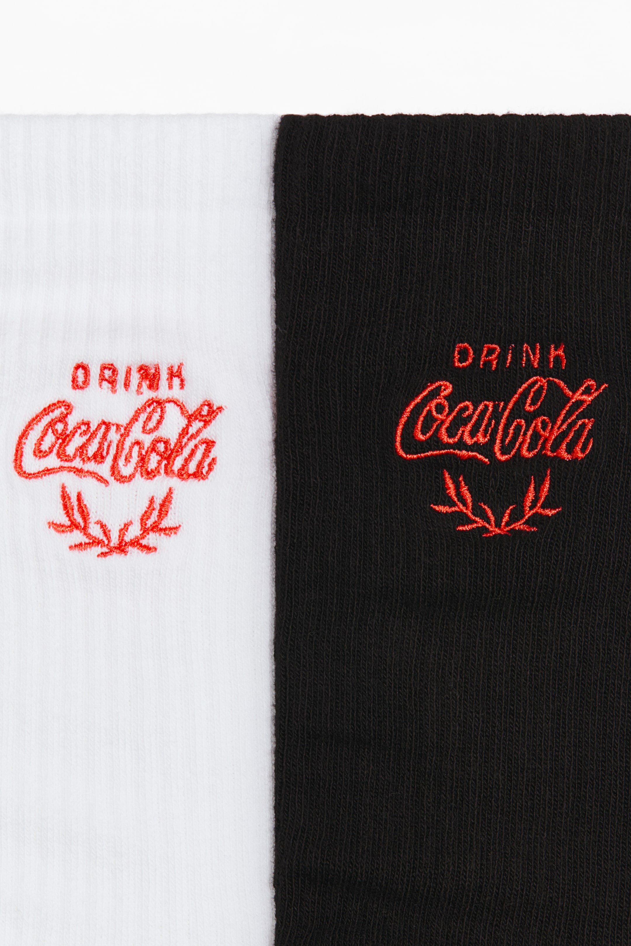 View larger image: A pair of socks, crafted from a soft, fine-knit cotton blend, is being displayed. One sock is white and the other is black, and each high, ribbed shaft features a red embroidered 'DRINK Coca-Cola' logo with a stylized leaf motif beneath it.