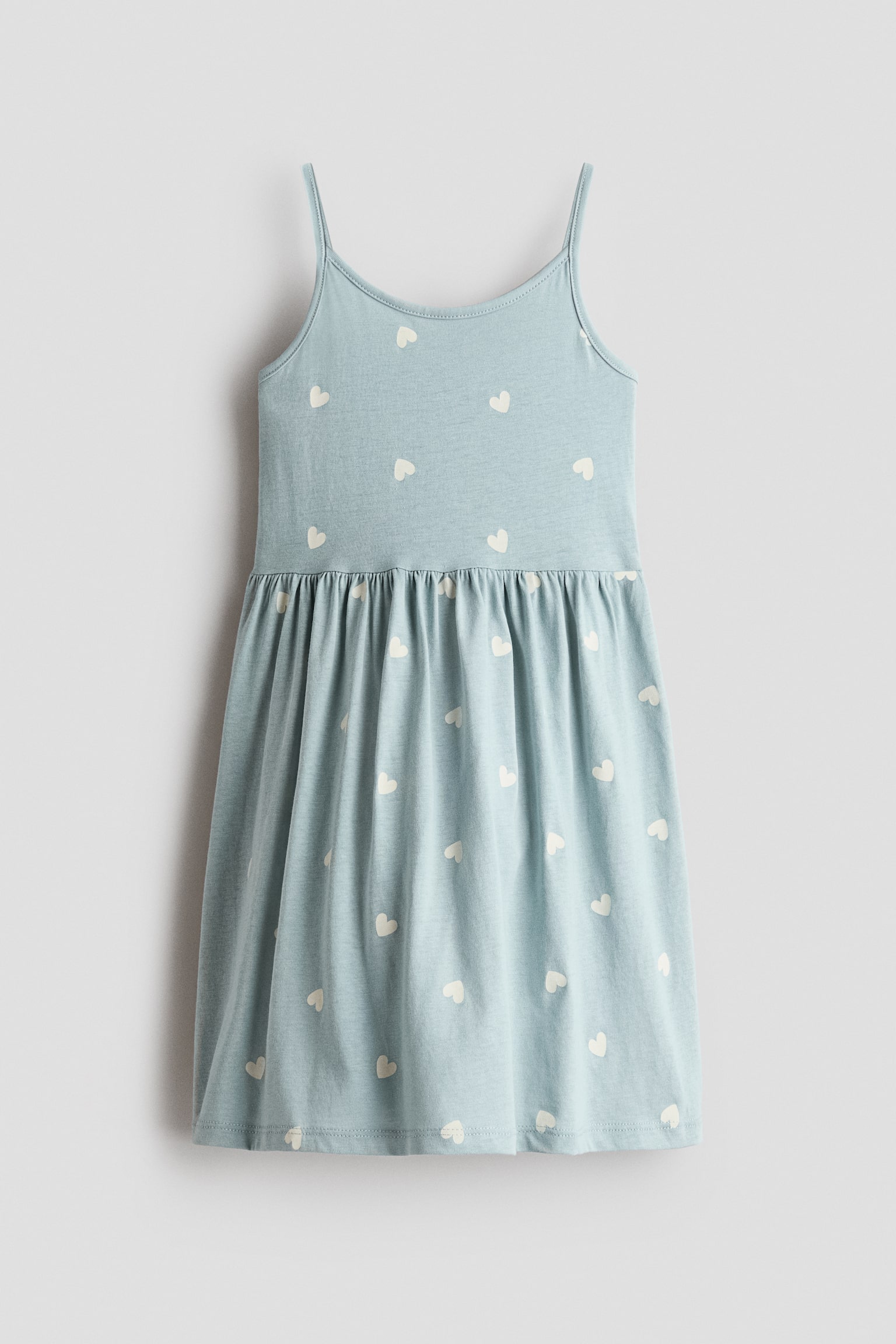 A light blue cami dress is shown from the front, featuring thin spaghetti straps, a scoop neckline, and an all-over pattern of small cream-coloured hearts. The dress showcases a gathered waistline that flows into a flared skirt.
