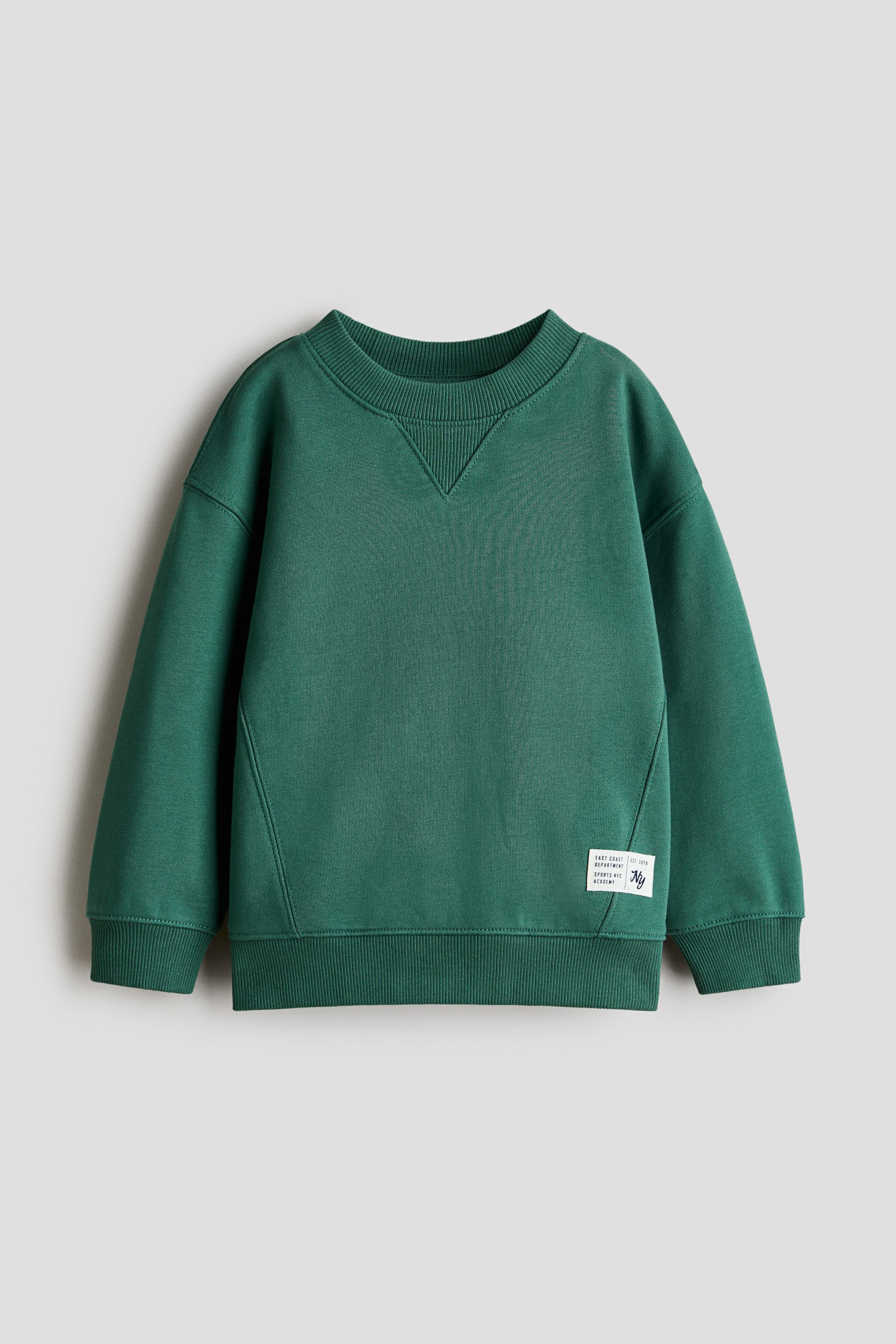 Se større billede: Bomulds sweatshirt præsenterer en frontvisning, der fremhæver en mørkegrøn farve, en ribbet rund hals med V-søm detalje under halsudskæringen og lange ærmer med ribbede manchetter. Den er designet med en ribbet kant, diagonale sømme tværs over den nedre frontdel og et rektangulært logomærke på den nederste højre side.