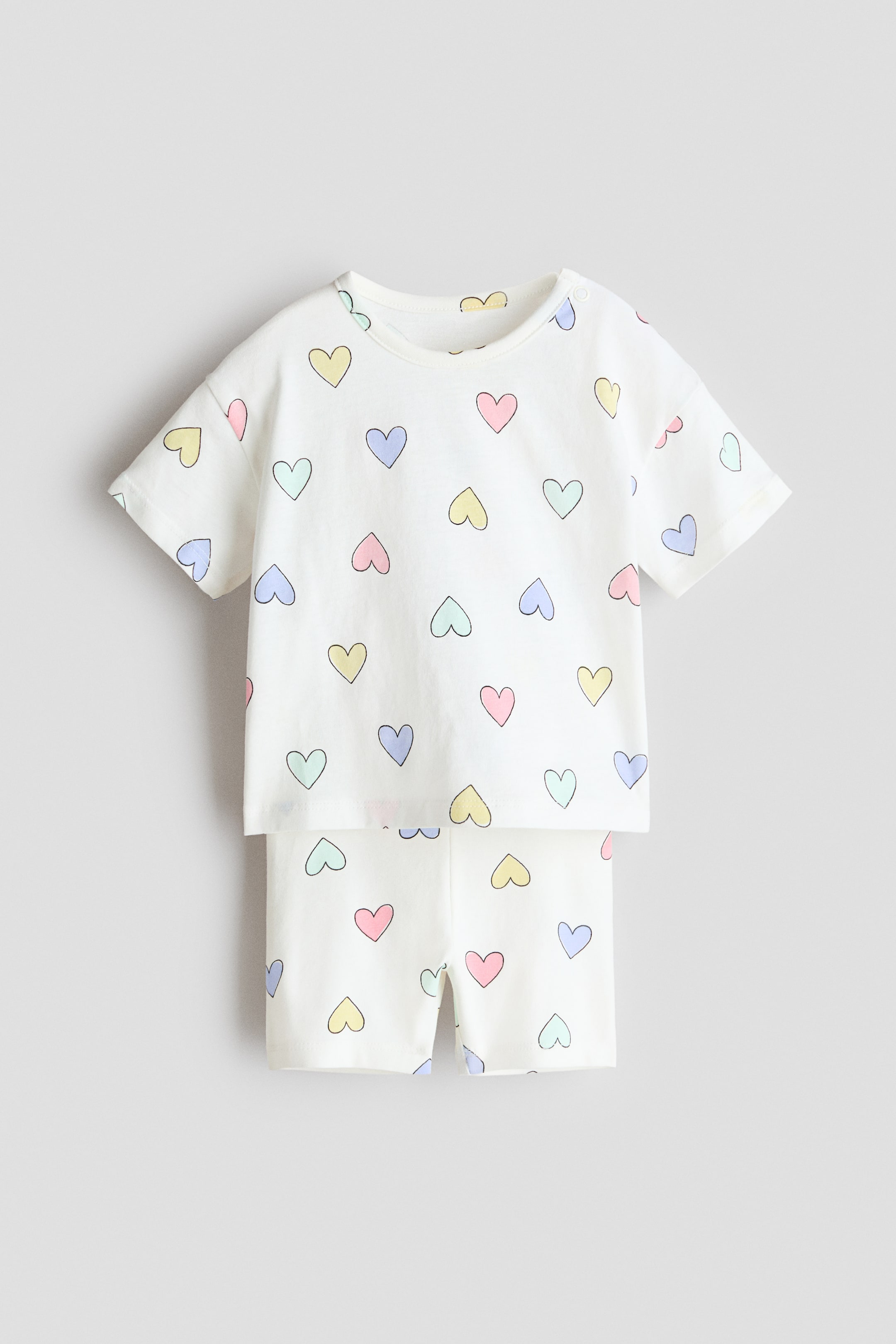 Büyük resmi görüntüle: Beyaz bebek pijama takımı, pastel kalp desenleriyle süslenmiş kısa kollu bir üst ve uyumlu şortlardan oluşmaktadır. Üst, bir omzunda çıtçıtlı yuvarlak bir yaka ile sunulurken, şort kısa kesimiyle dikkat çekiyor.