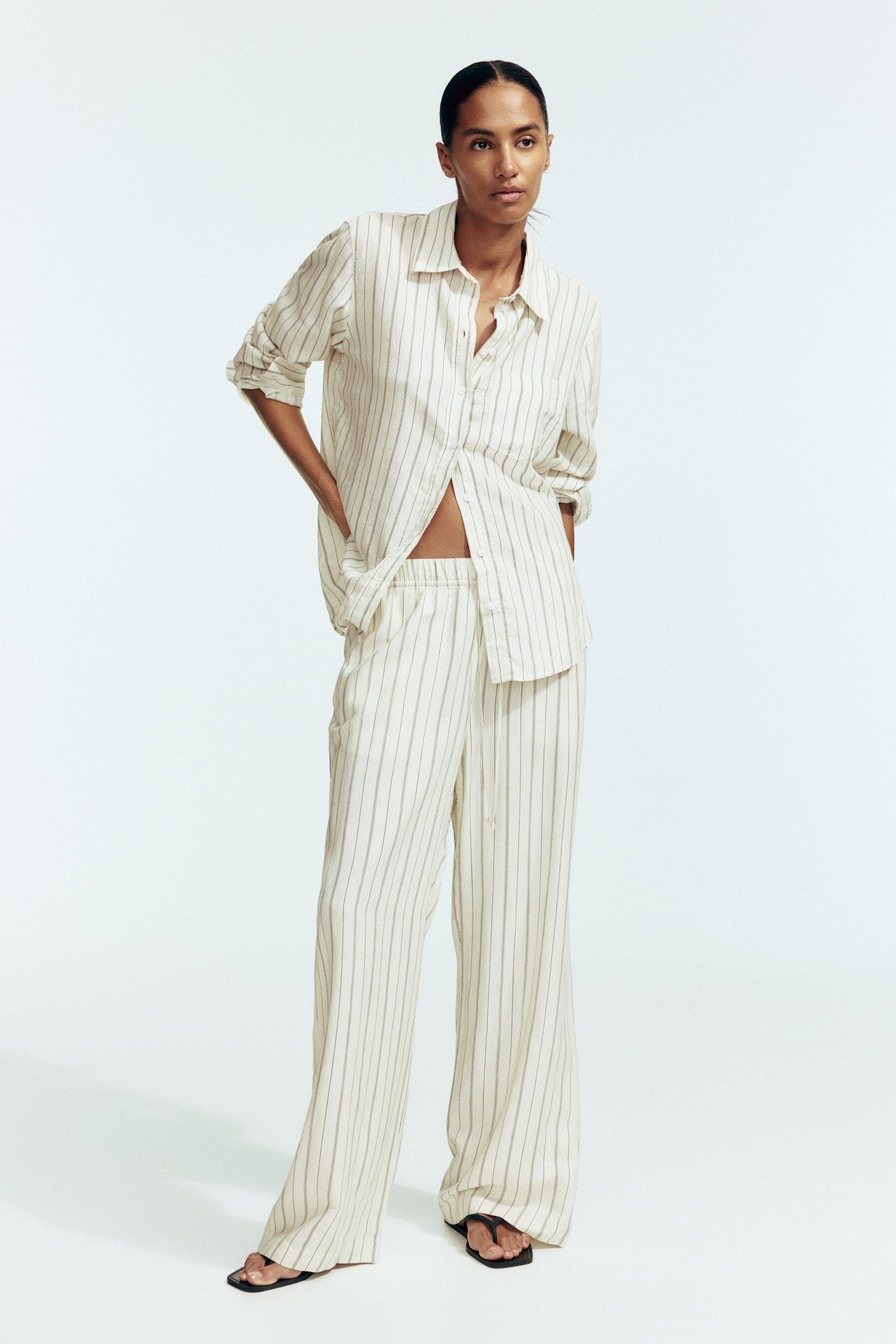 View larger image: An off-white two-piece set is being presented, consisting of a collared shirt and wide-leg pants, both featuring thin vertical stripes. The shirt is showcasing rolled-up long sleeves and a chest pocket, while the pants are featuring an elasticated drawstring waistband and a flowing silhouette.