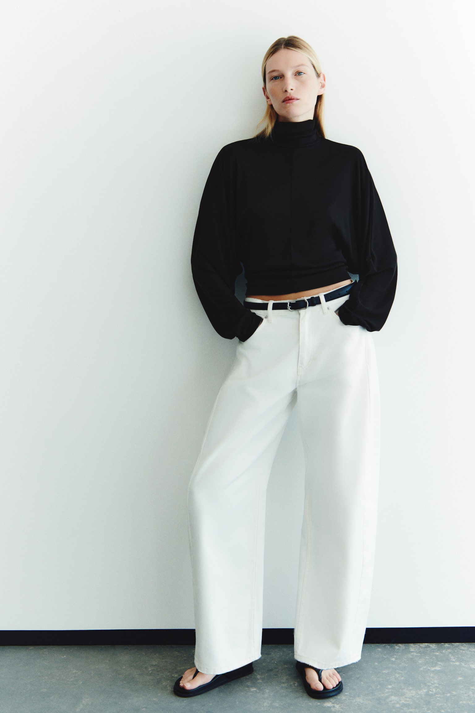 A black, fitted knit top featuring a double-layered high neck, long batwing sleeves, and a visible centre-front seam is being worn by the model. The top is styled with white wide-leg trousers and a dark belt. - 1
