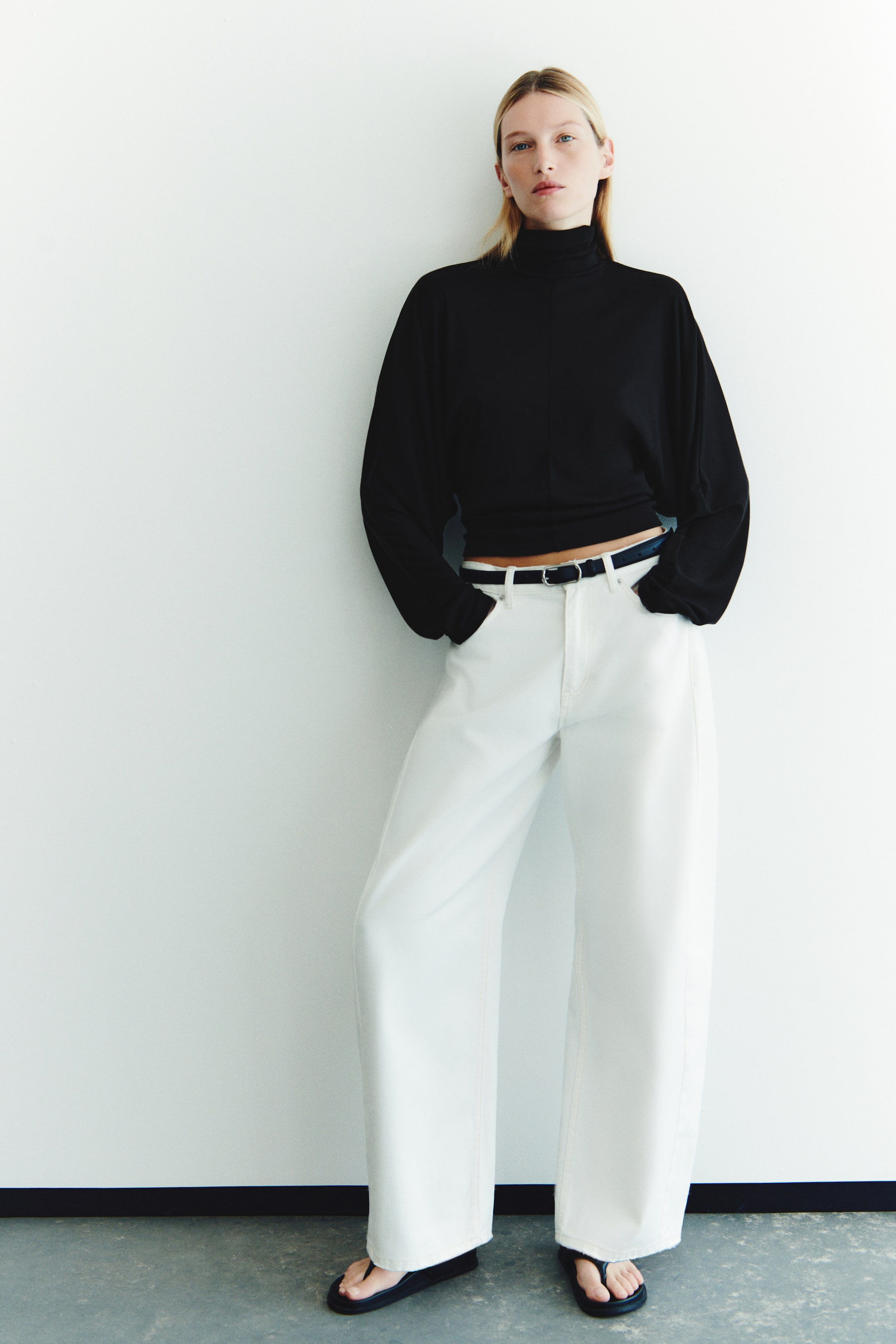 View larger image: A black, fitted knit top featuring a double-layered high neck, long batwing sleeves, and a visible center front seam is being worn by the model. The top is styled with white wide-leg trousers and a dark belt.