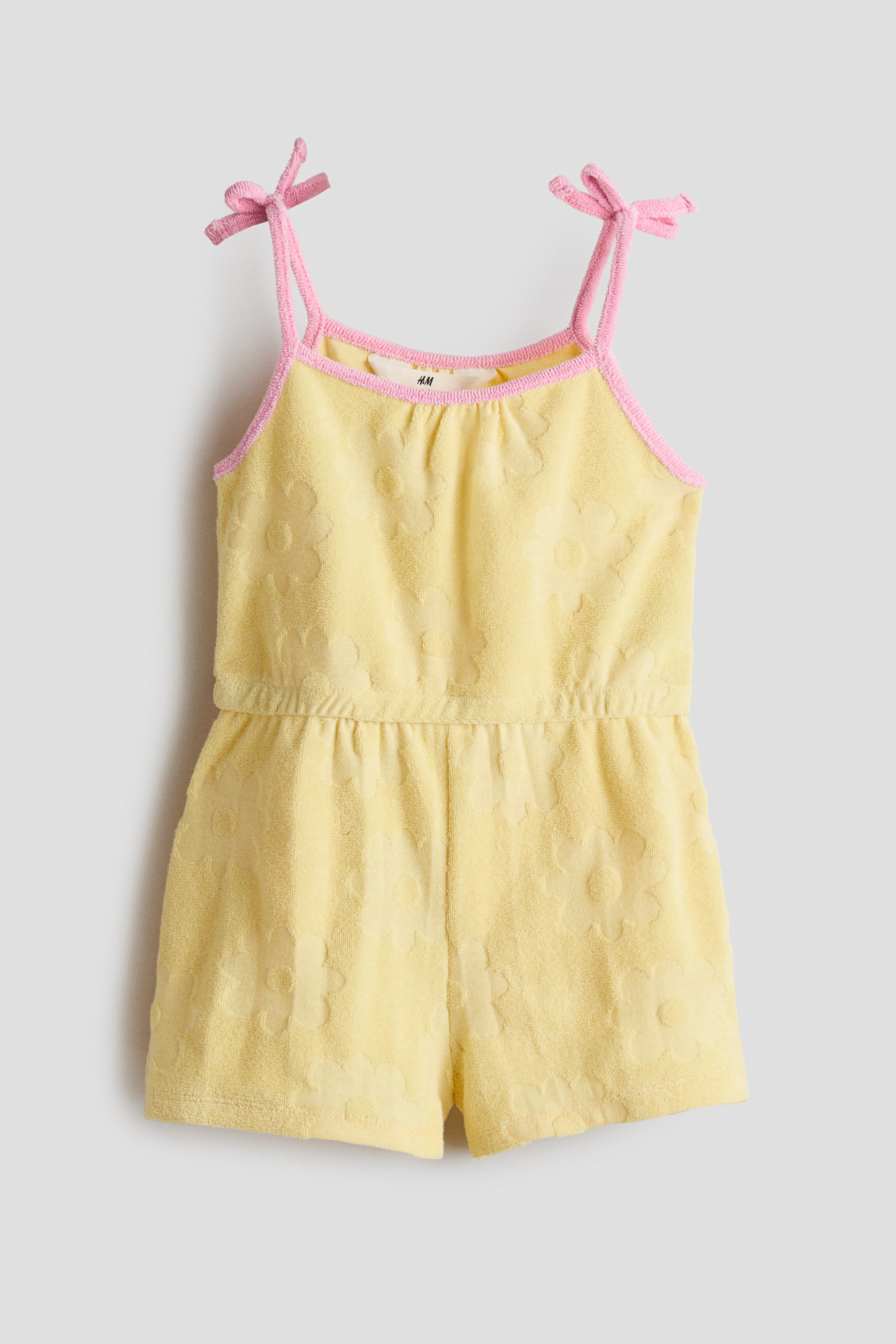 Größeres Bild ansehen: Ein gelber, weicher Frottee-Jumpsuit wird von vorne präsentiert, mit einem geprägten Blumenmuster, einem geraden Ausschnitt mit rosa Paspeln und schmalen Schulterträgern, die sich zu rosa Schleifen binden. Eine verdeckte Elastik sorgt für eine taillierte Passform.