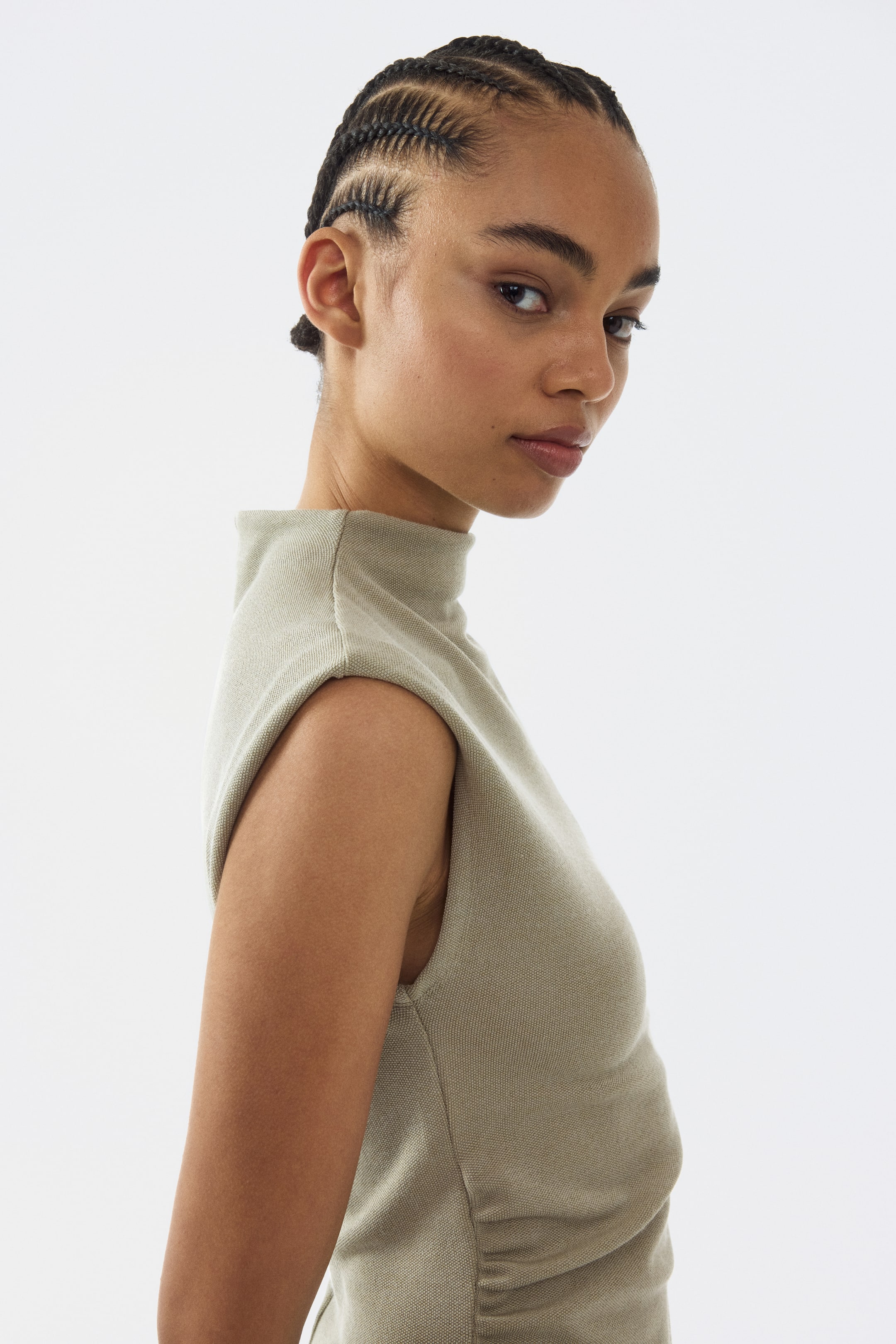 Prikaži veću sliku: Svetlo maslinasto zeleni top bez rukava prikazuje fino teksturiranu pletenu tkaninu. Odlikuje ga lažni okovratnik i produžene linije ramena, sa suptilnim nabiranjem vidljivim duž bočnog šava, predstavljajući usku siluetu iz ugla od tri četvrtine.