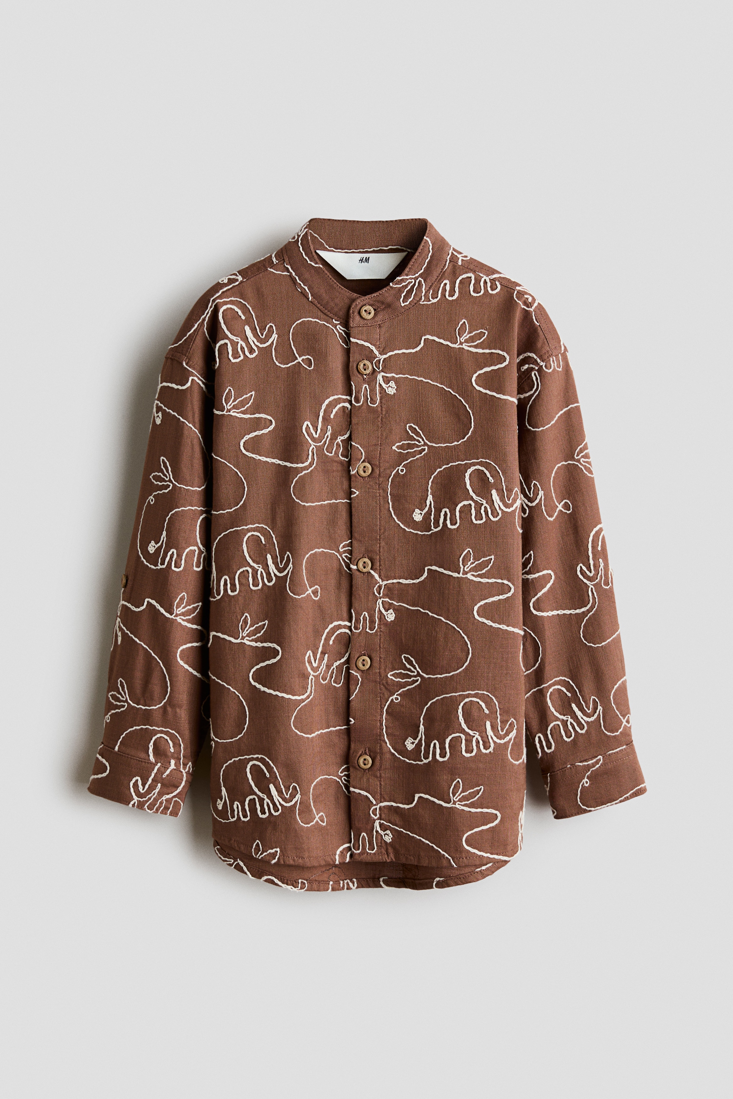 A brown grandad shirt featuring an all-over white continuous-line elephant pattern is shown from the front, showcasing a band collar, a button-down front, and long sleeves with rolled-up cuffs.