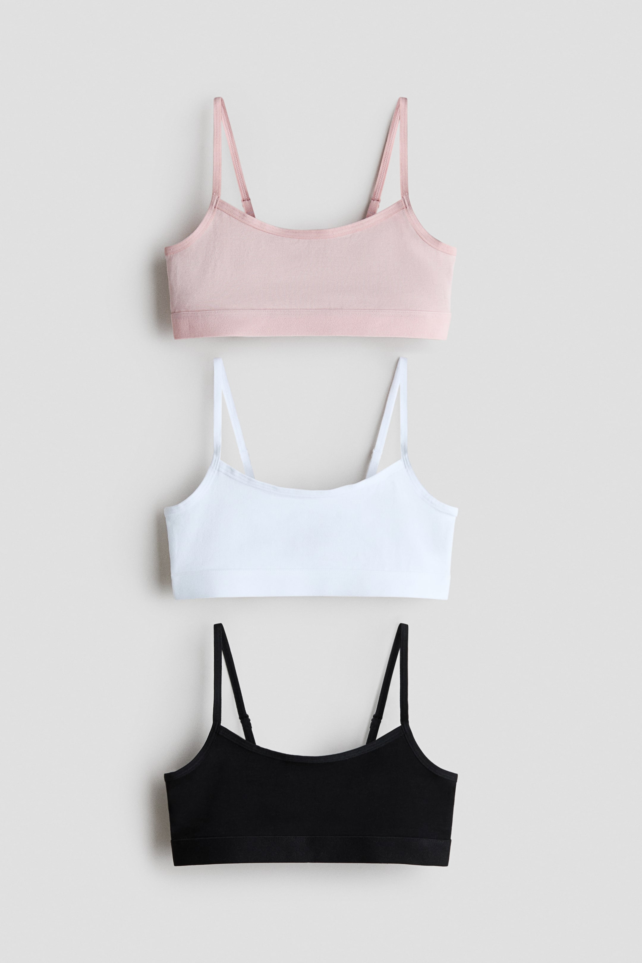 Grotere afbeelding bekijken: Drie crop tops van zachte katoenen jersey liggen plat en worden van voren getoond. Ze zijn afgebeeld in roze, wit en zwart, elk met smalle, verstelbare schouderbandjes en een elastische zoom.