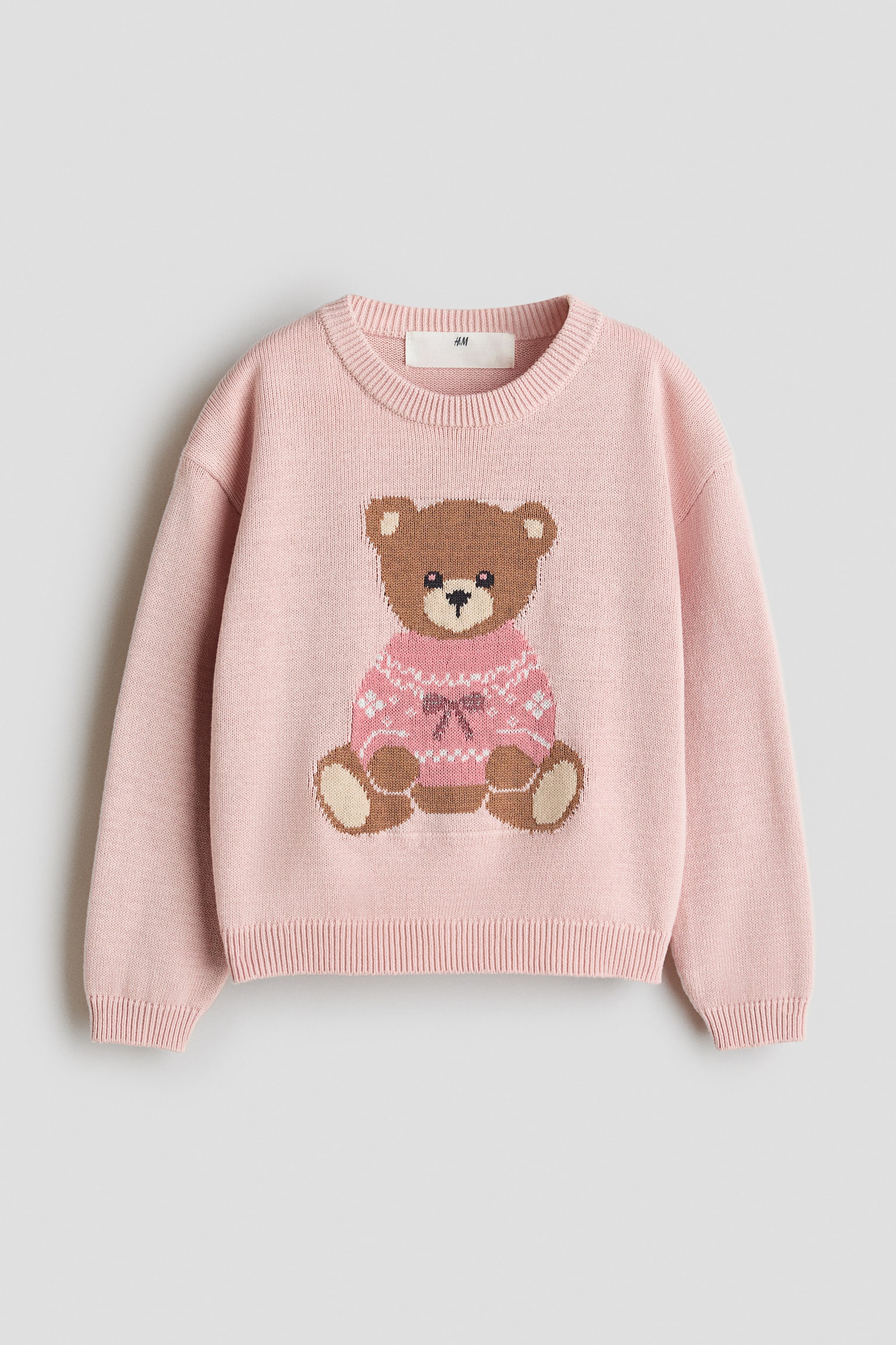 Se muestra de frente un jersey de algodÃ³n de punto jacquard rosa, con un grÃ¡fico central de un oso de peluche marrÃ³n que lleva un jersey rosa estampado. El diseÃ±o incluye un cuello redondo con acabado acanalado, hombros caÃ­dos, mangas largas con puÃ±os acanalados y bajo acanalado.