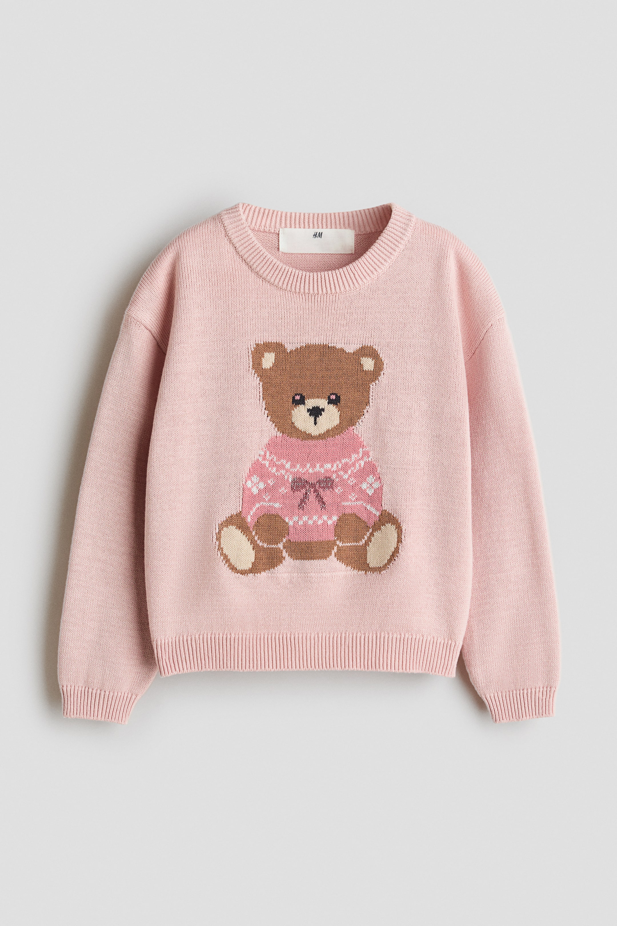 Agrandir l'image: Un pull en coton tricoté jacquard rose est présenté de face, orné au centre d’un graphisme représentant un ours en peluche brun portant un pull rose à motif. Le modèle présente un col rond bordé de côtes, des épaules tombantes, des manches longues avec poignets côtelés et un ourlet côtelé.