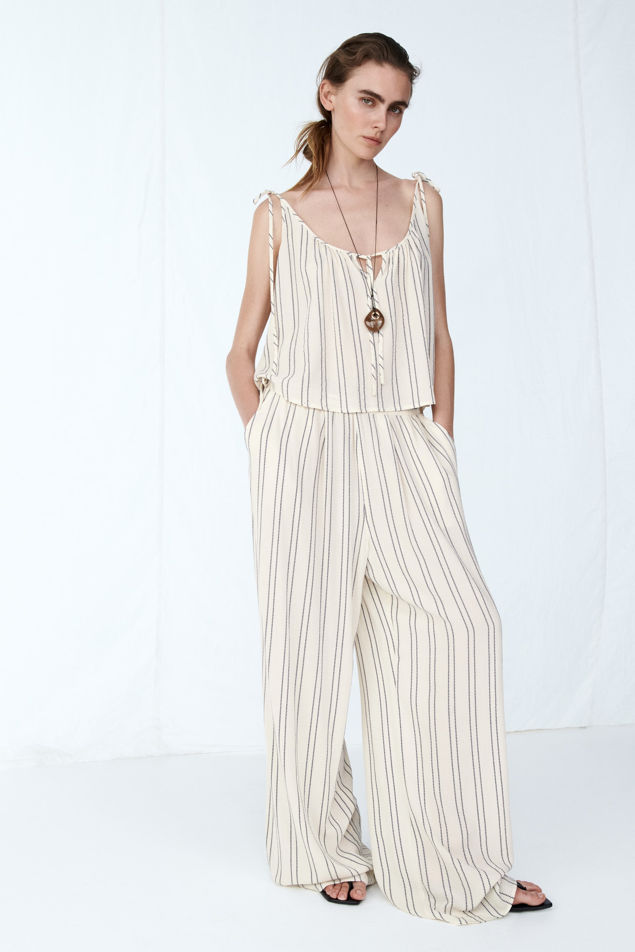 View larger image: This two-piece set highlights a sleeveless camisole top with adjustable shoulder ties and a V-neckline, complemented by matching wide-leg pants. Both pieces feature vertical dark pinstripes on a light-toned fabric.