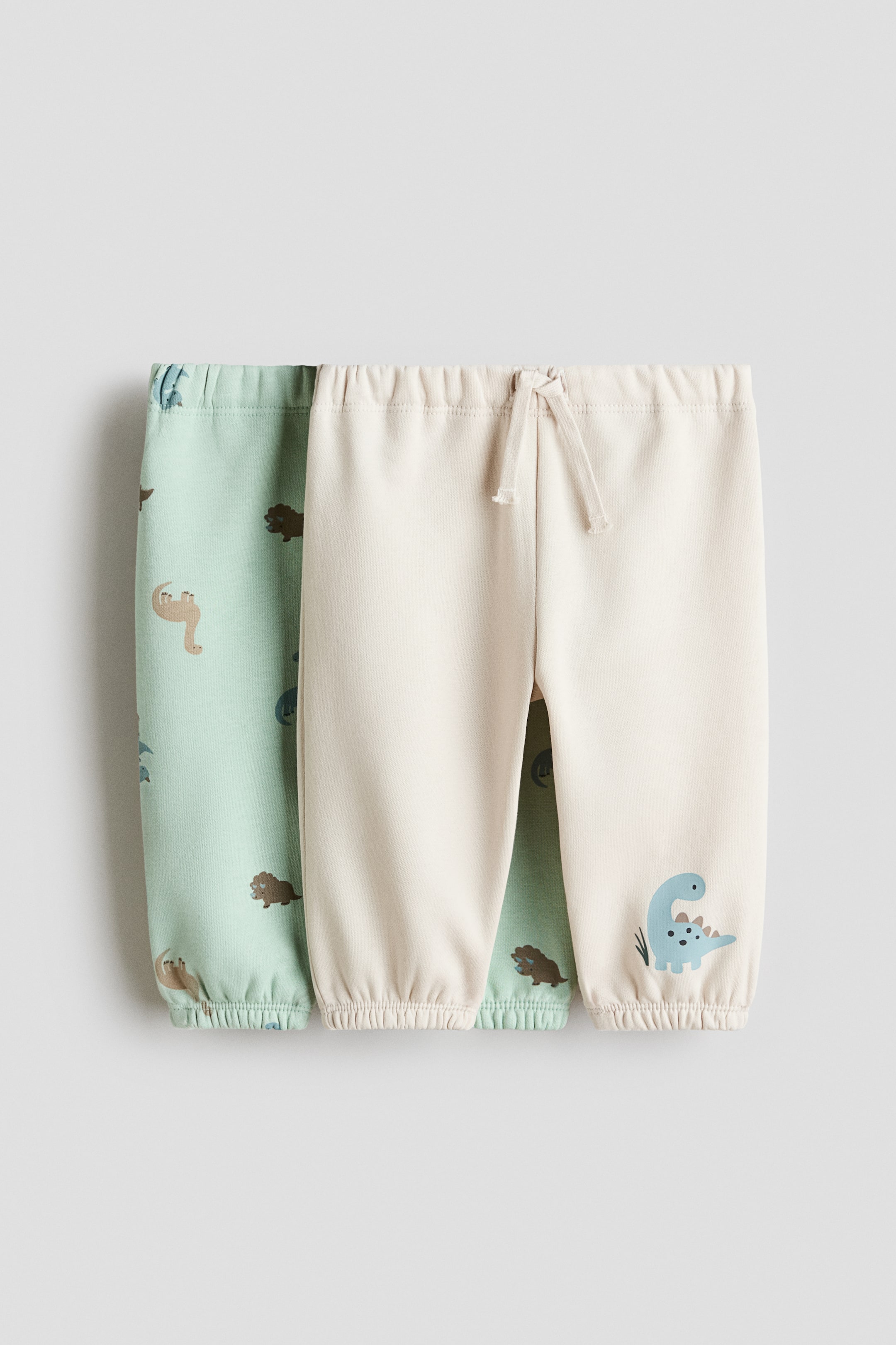 View larger image: Two pairs of baby trousers are shown from the front. One light-green pair features an all-over dinosaur print and elasticated cuffs, while the other light-beige pair has an elasticated drawstring waistband, elasticated cuffs and a blue dinosaur graphic near the left ankle.