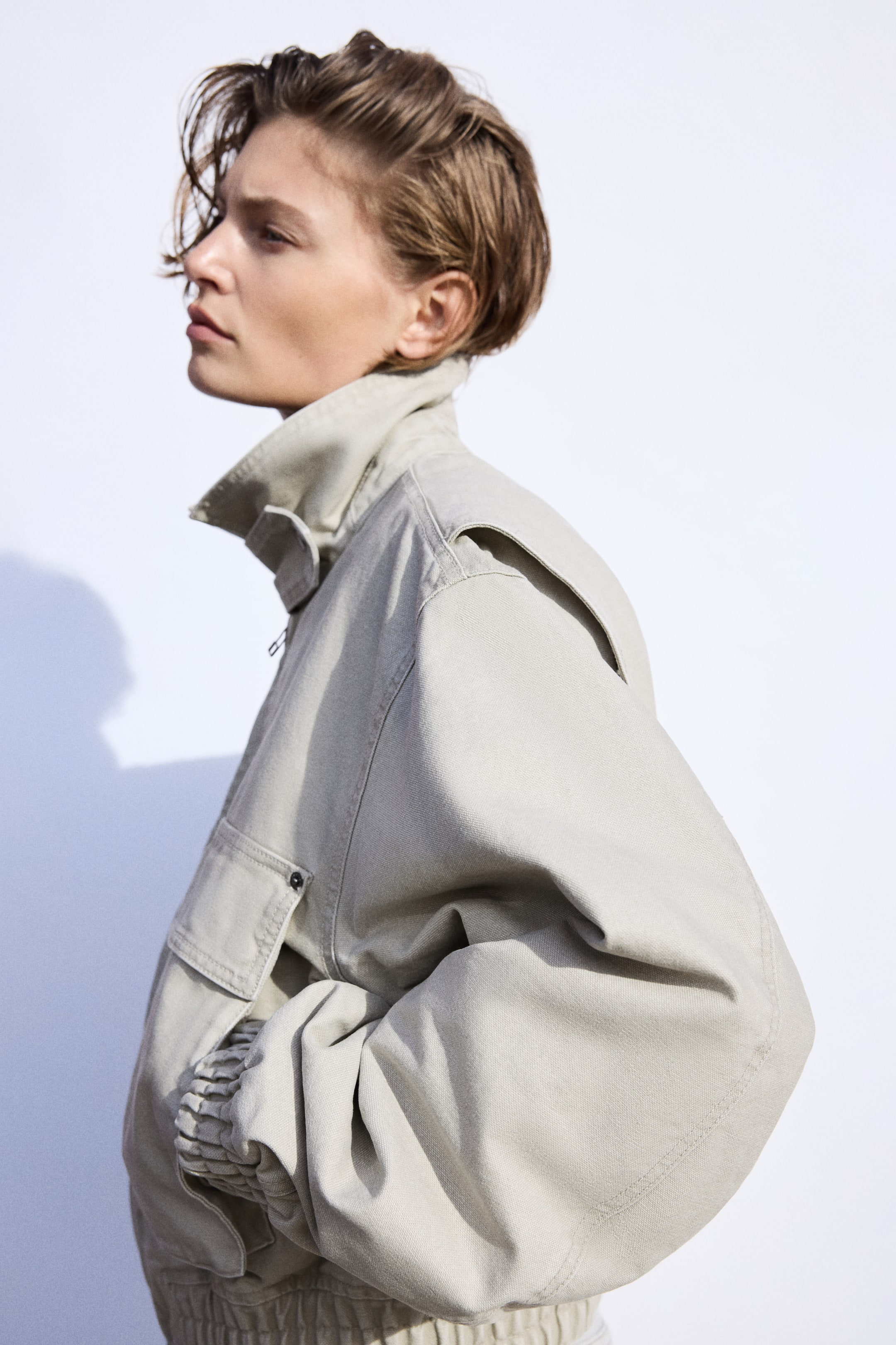 拡大画像を表示: ライトベージュのボンバージャケットは、横向きのシルエットから高めのスタンドカラー、ドロップショルダー、リブカフス付きのボリュームスリーブを見せています。左袖にはカーゴポケットのディテールがあります。