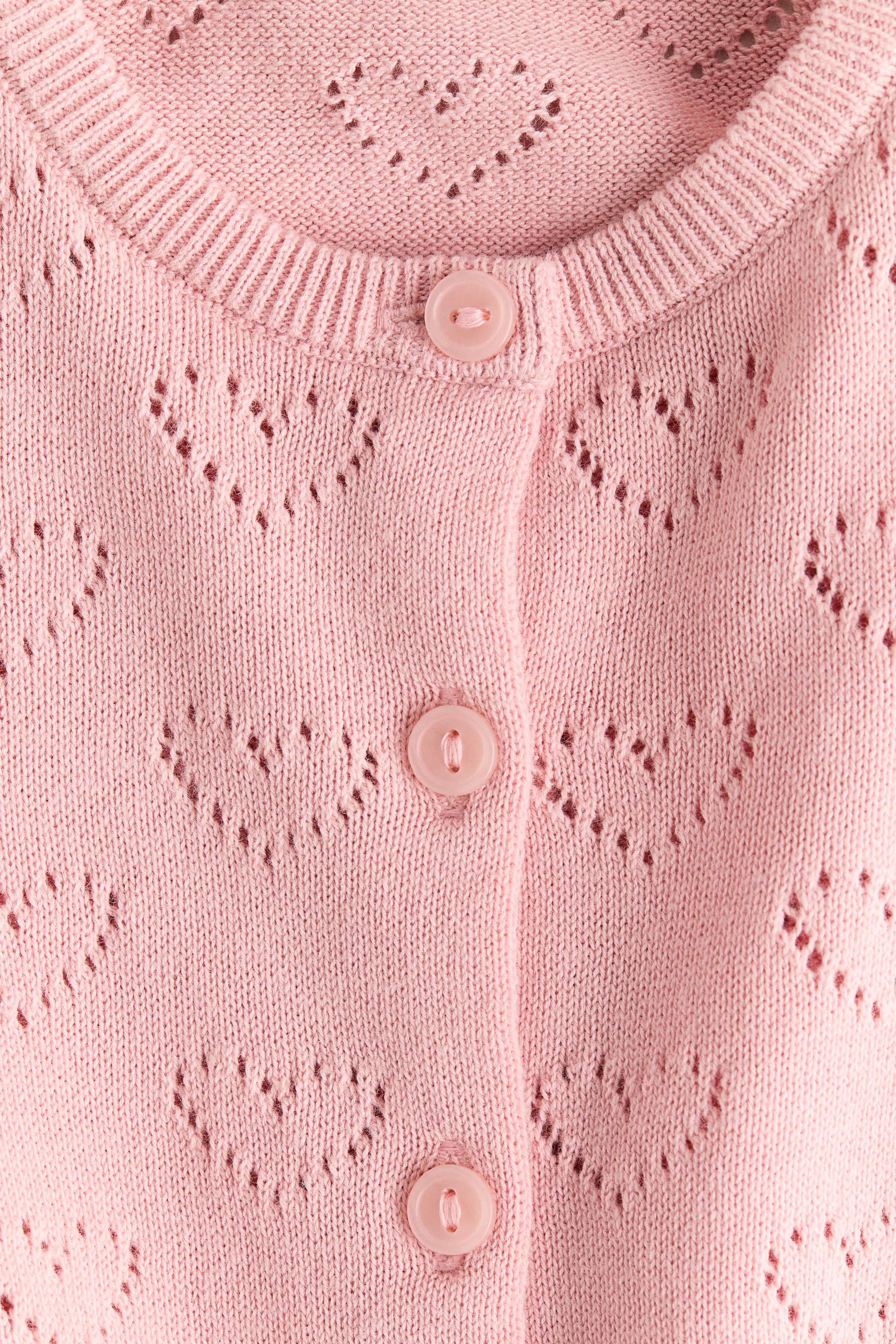 Grösseres Bild ansehen: Ein rosa Cardigan wird in einer VORDERANSICHT präsentiert, mit einem Musterstrick und offenen herzförmigen Details, einem runden gerippten Ausschnitt und drei passenden rosa Knöpfen, die entlang der vorderen Knopfleiste befestigt sind.