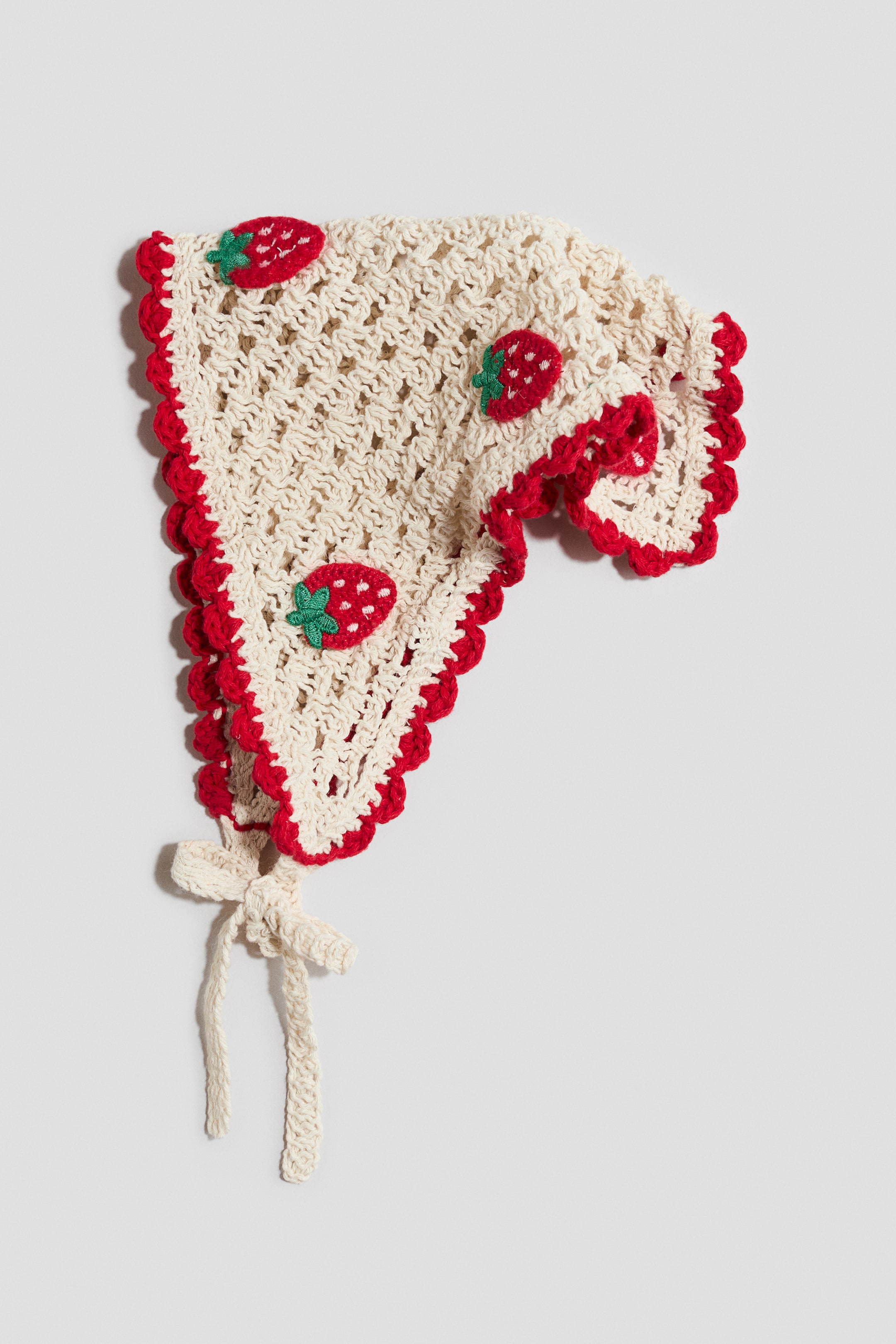 View larger image: A crochet bonnet features a cream-colored, open-weave pattern with red scalloped trim along its edges. Small red strawberries with green leaves are stitched onto the fabric, and cream-colored crochet ties are knotted into a bow at the bottom.