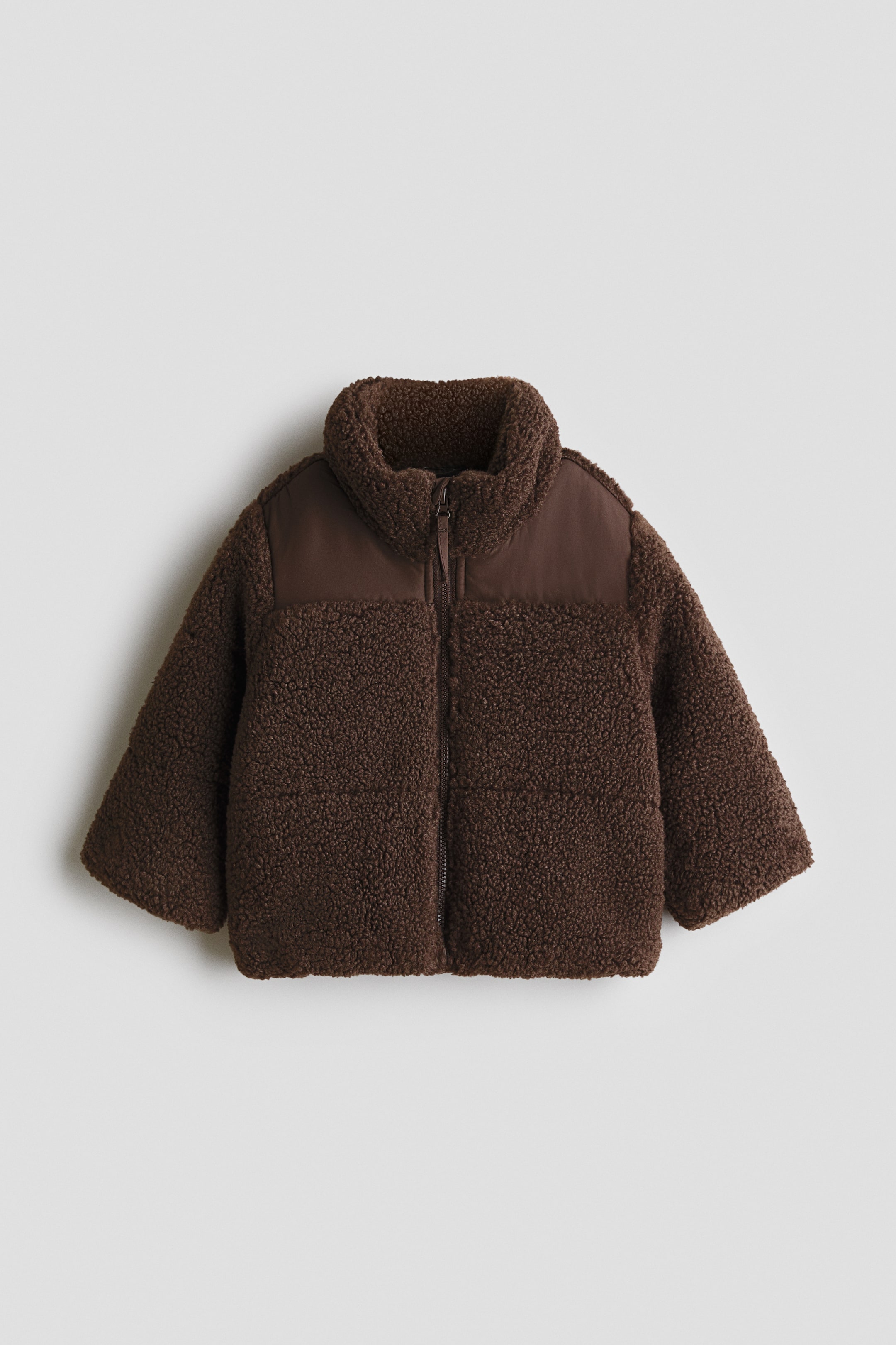 Größeres Bild anzeigen: Eine dunkelbraune Jacke ist von vorne zu sehen. Sie verfügt über eine Teddyfleece-Struktur an Body und Ärmeln, die durch glatte, dunkelbraune Einsätze an den Schultern und einen Stehkragen kontrastiert wird. Ein Frontreißverschluss verläuft mittig entlang der Vorderseite.