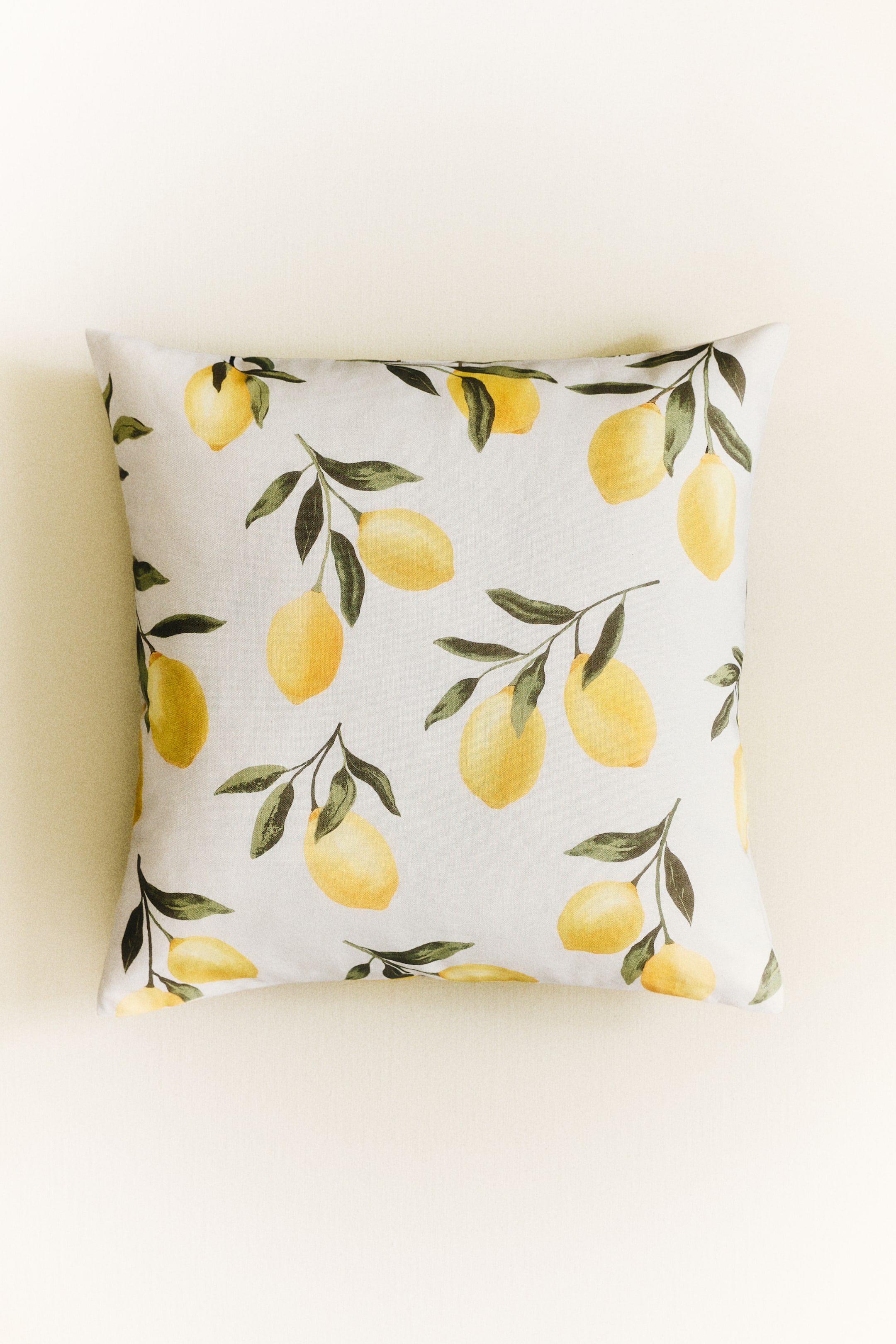 View larger image: A square cushion is displayed horizontally, featuring a repeating pattern of yellow lemons on branches with green leaves against a light grey fabric.