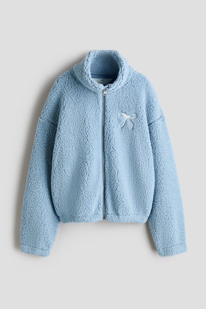 Una giacca teddy in pile azzurro è mostrata frontalmente, con colletto alto alla coreana, zip frontale integrale e spalle scese. La giacca dalla vestibilità morbida presenta polsini e fondo elasticizzati, oltre a un piccolo dettaglio a fiocco bianco sul petto sinistro. - 1