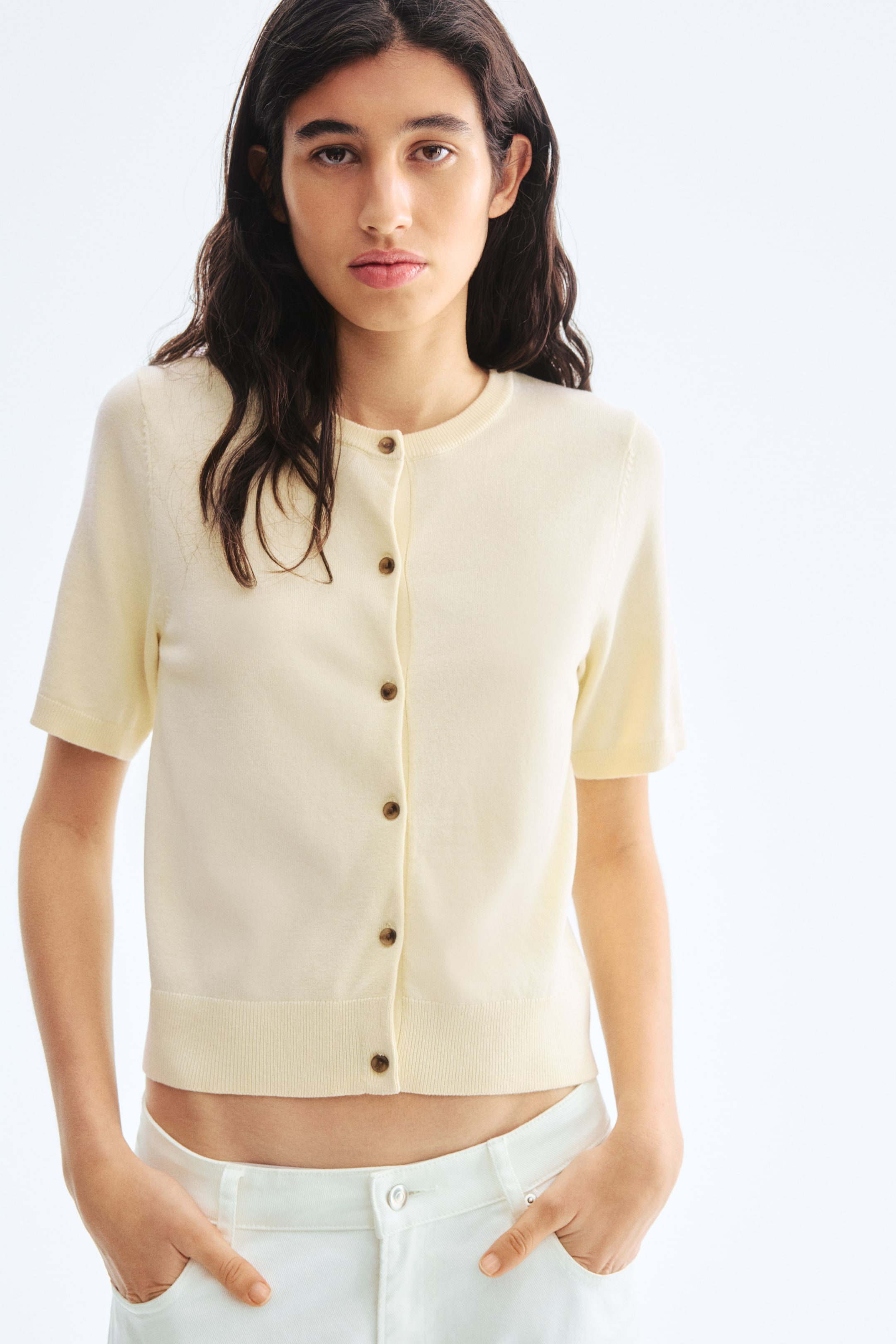 View larger image: A light yellow short-sleeved cardigan, made of a fine knit, is being worn by the model. The cardigan features a round, rib-trimmed neckline, brown buttons down the front, and ribbing at the cuffs and hem, and is being styled with white pants.
