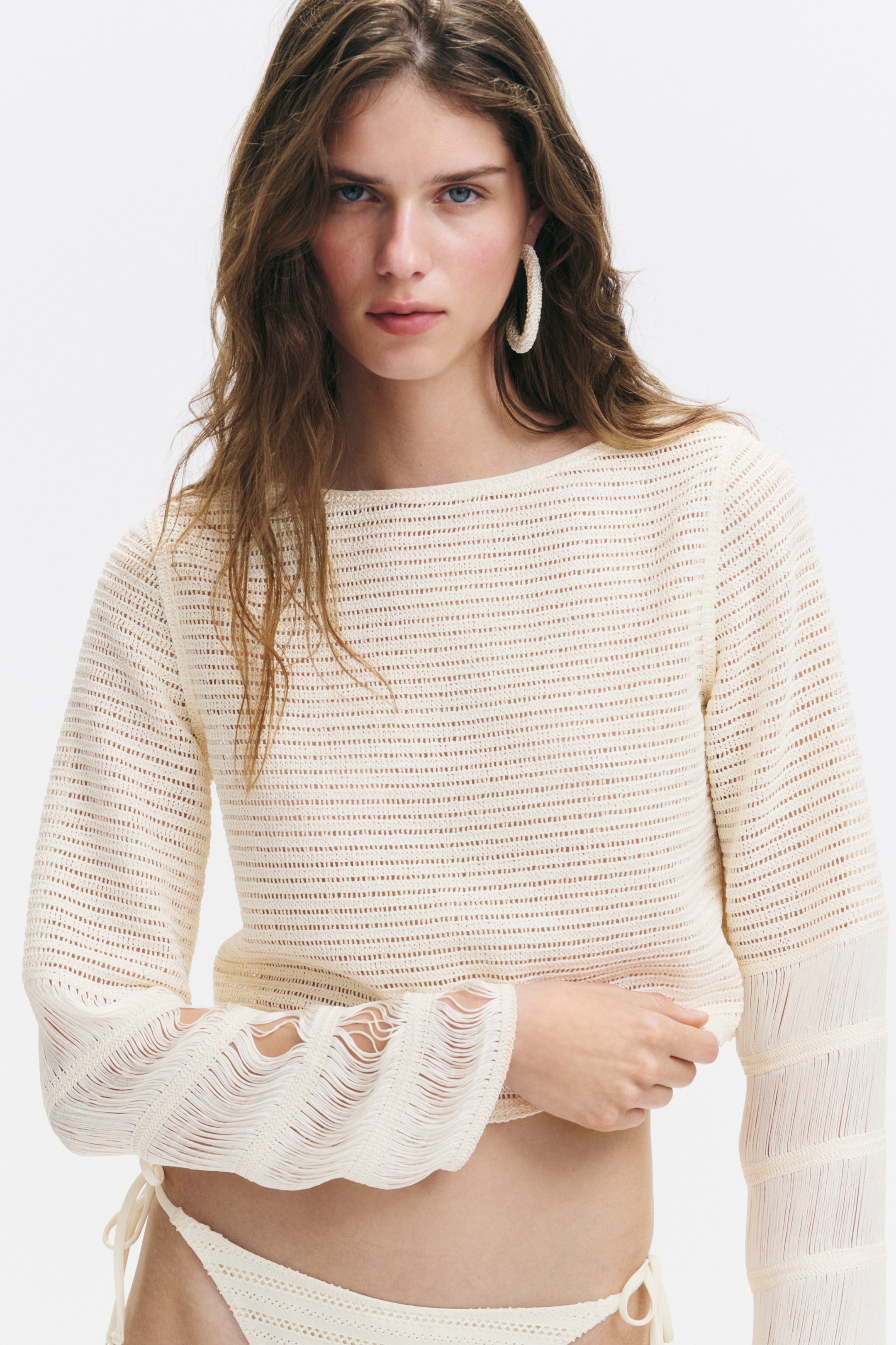 View larger image: A cream open-knit cropped top with a boat neck and long sleeves featuring fringed cuffs is being worn, paired with matching open-knit bikini bottoms with side ties.