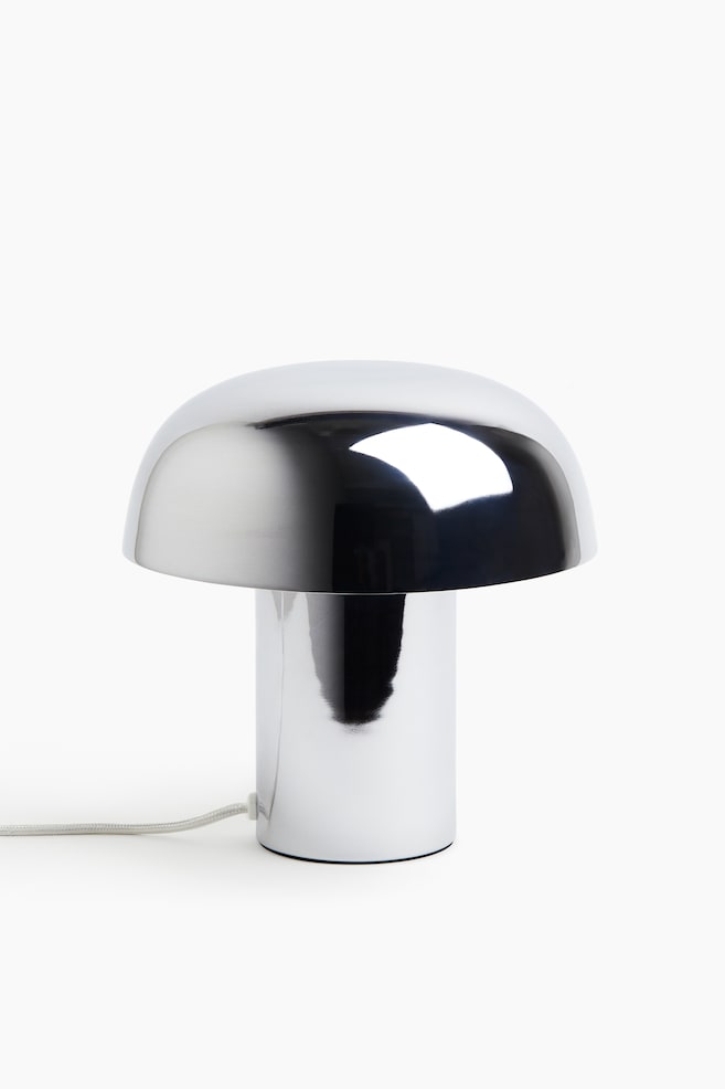 Eine moderne, pilzförmige Tischlampe mit hochreflektierender Chromoberfläche steht frontal ausgerichtet. Die Lampe zeigt einen breiten, abgerundeten Kuppelschirm, der auf einem dicken zylindrischen Sockel ruht, mit einem hellgrauen, stoffummantelten Kabel, das aus der Unterseite herausführt. - 1