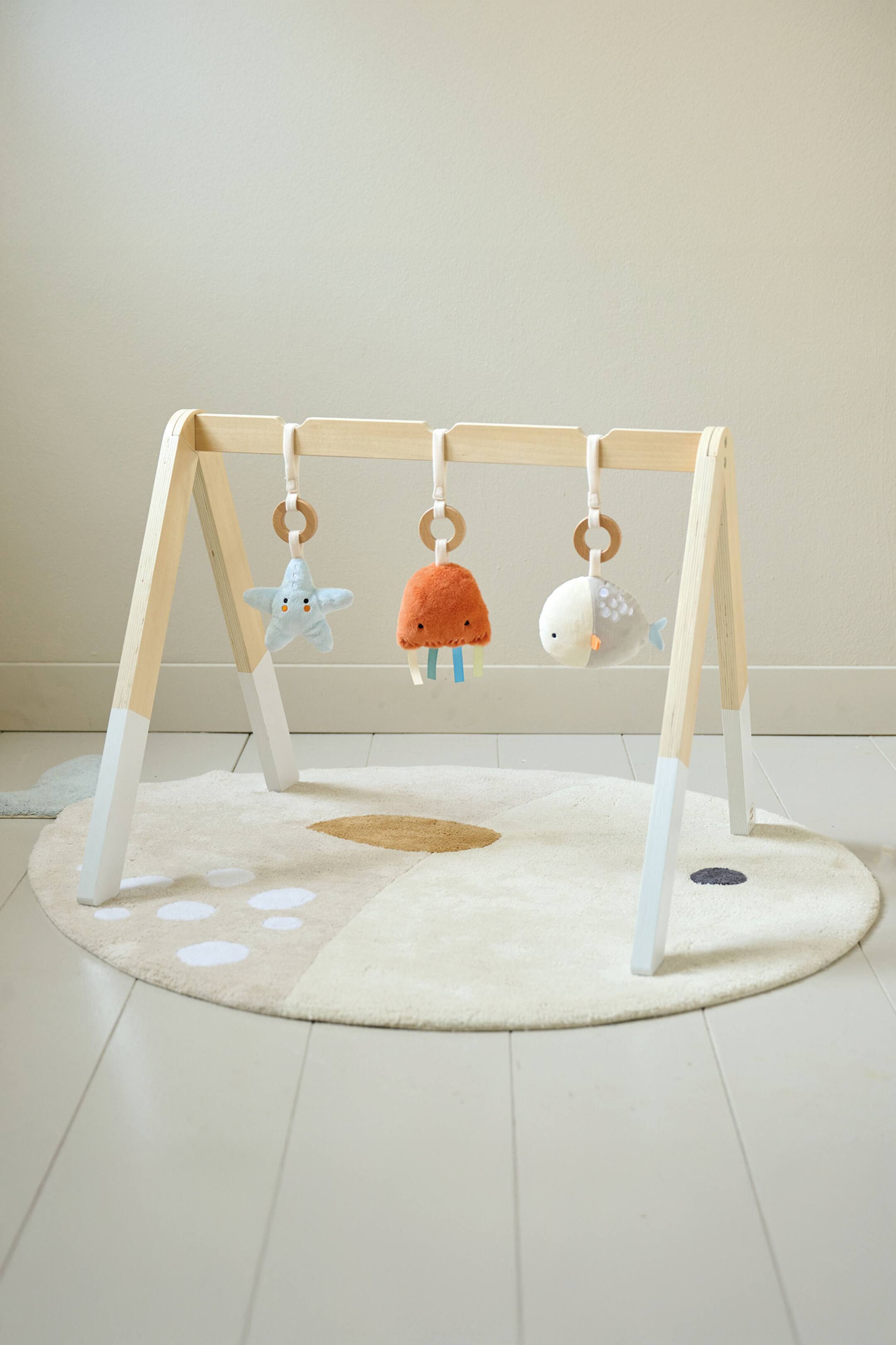 Grotere afbeelding bekijken: Een houten baby speelgymnastiek, met natuurlijke houten A-frame poten met witgedompelde bases, staat vooraan op een ronde, lichtgekleurde mat. Drie zachte speelgoedjes, een blauwe zeester, een oranje kwal met kleurrijke stroken, en een grijze walvis, hangen aan de horizontale balk door middel van stoffen lussen en houten ringen.