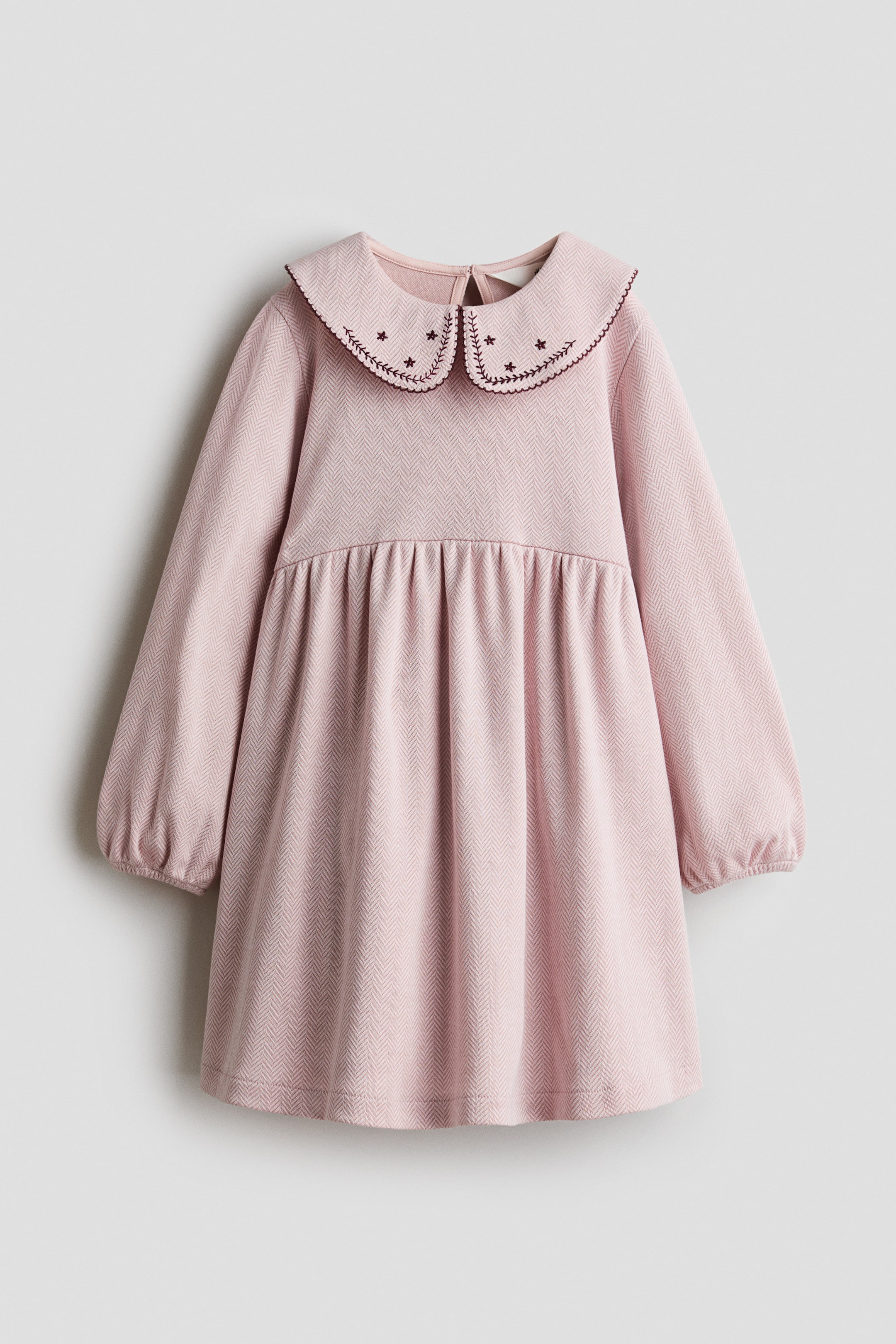 View larger image: A light pink long-sleeved dress in a textured herringbone jacquard jersey is shown facing front, featuring a rounded Peter Pan collar with dark red embroidered star motifs and scalloped edge-stitching. The dress displays a high gathered waistline that flows into a flared skirt, along with gathered elasticated cuffs.
