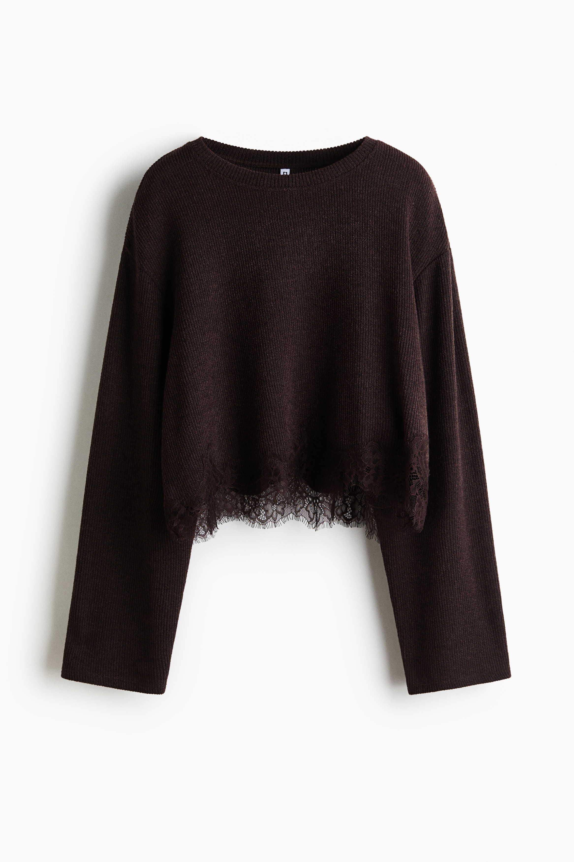 A short, loose-fit dark brown knit top featuring a round neckline, dropped shoulders and a delicate lace-trimmed hem is worn by the model. The model styles the top with dark fitted trousers, raising one arm to showcase the cropped length and long, wide sleeves.