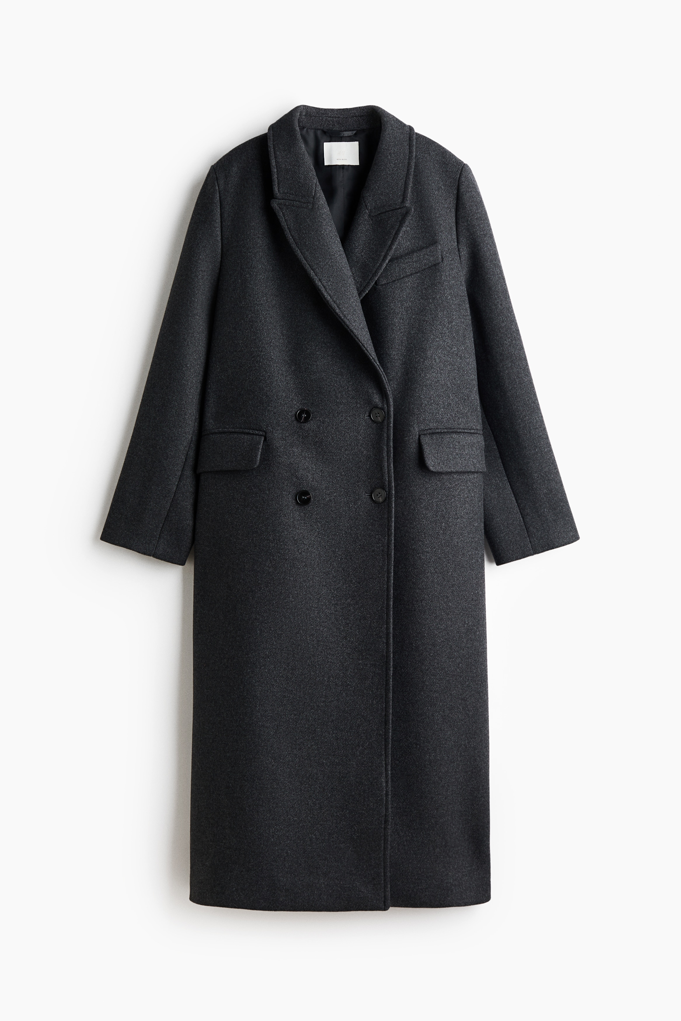A long, dark grey wool-blend coat with structured shoulders, peak lapels, and button cuffs is being worn by the model. The model is carrying a black leather handbag tucked under their arm while wearing black leather gloves.
