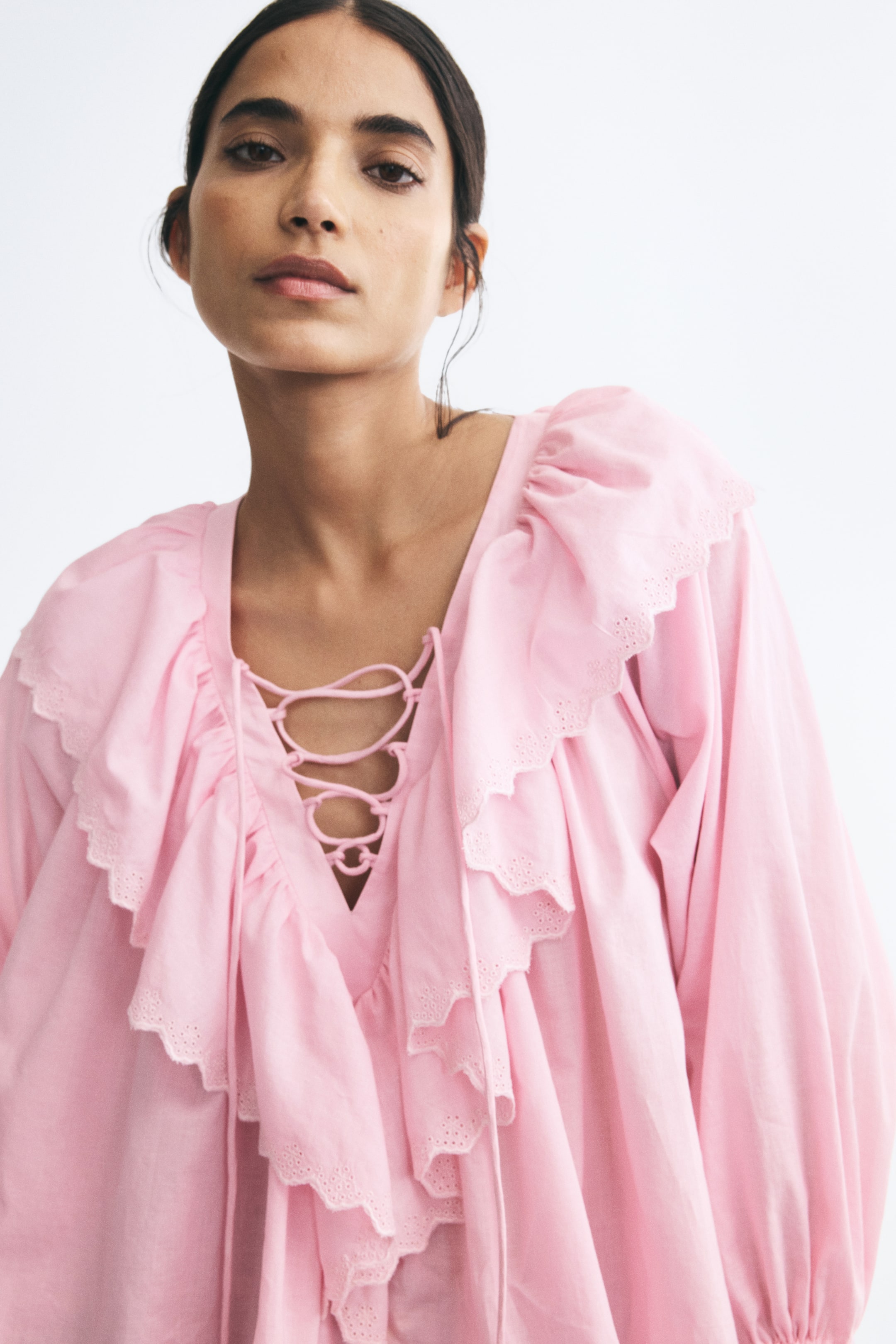 View larger image: The model is wearing a light pink, loose-fit blouse featuring a V-shaped neckline with a criss-cross lace-up front. The blouse is accented with a wide, scalloped ruffle trim, detailed with delicate broderie anglaise along the edges.