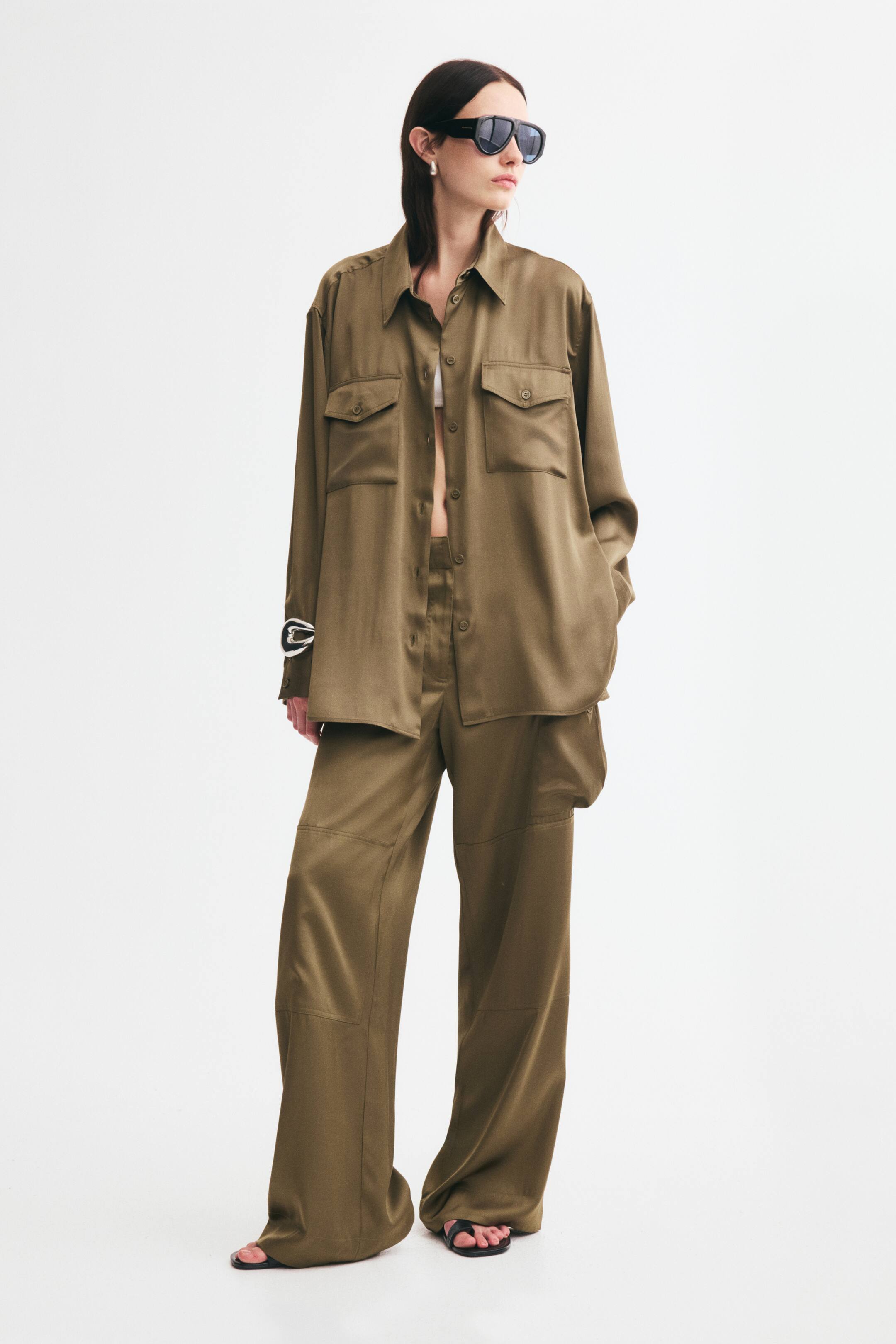 Xem ảnh lớn hơn: Một chiếc áo sơ mi satin màu xanh ô liu dáng oversized, có cổ áo, hàng cúc phía trước và túi ngực đắp nắp cài cúc, đang được người mẫu mặc mở cúc. Chiếc áo với độ rũ thoải mái và vai rơi nhẹ được phối cùng quần satin ống rộng đồng bộ và áo crop top trắng.