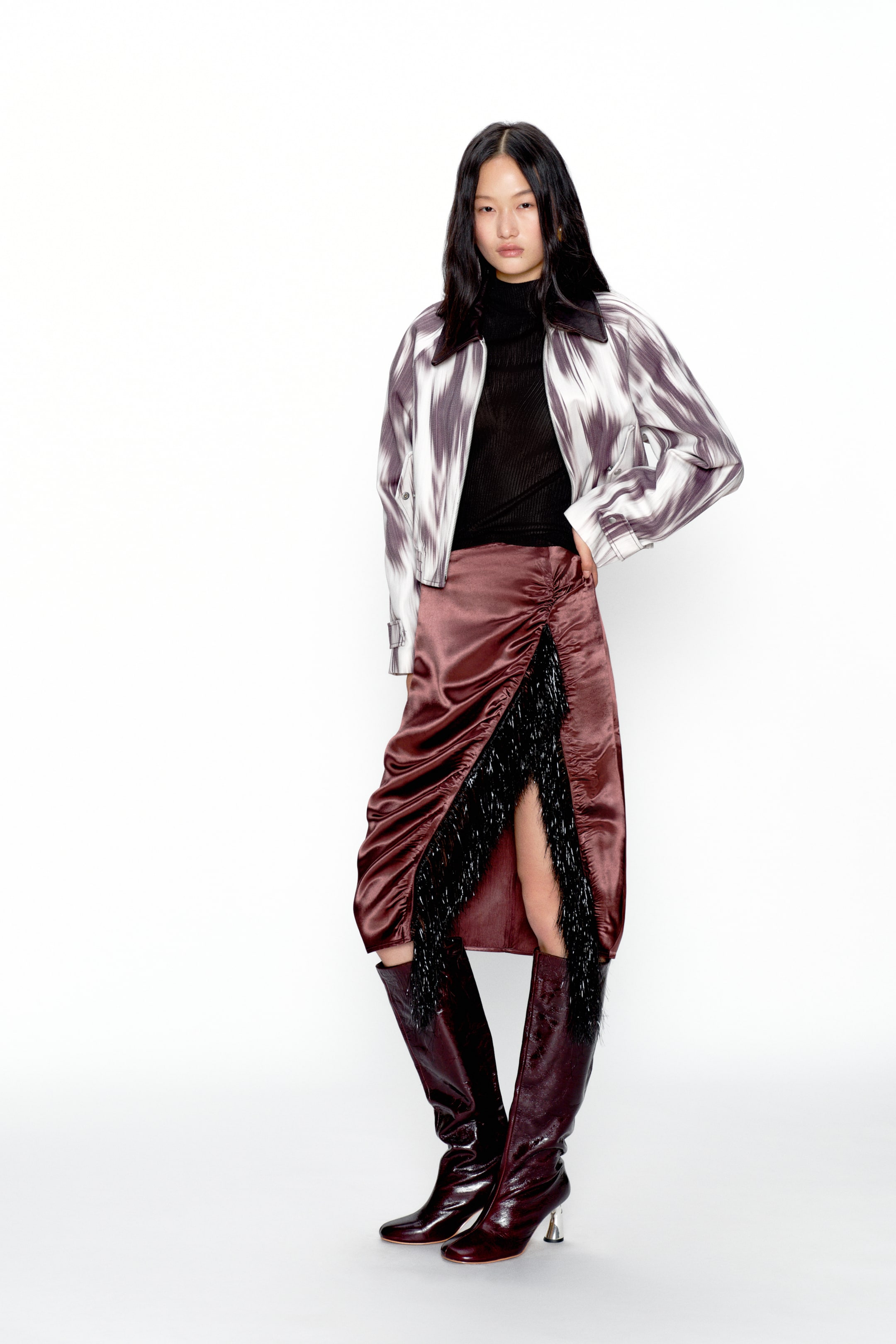 View larger image: A cropped jacket showcases a white and grey abstract pattern with a wide pointed collar, worn open over a black ribbed high-neck top. A reddish-brown satin midi skirt features heavy ruching and a high slit that reveals black fringe detailing, while matching knee-high boots complete the look with silver block heels.