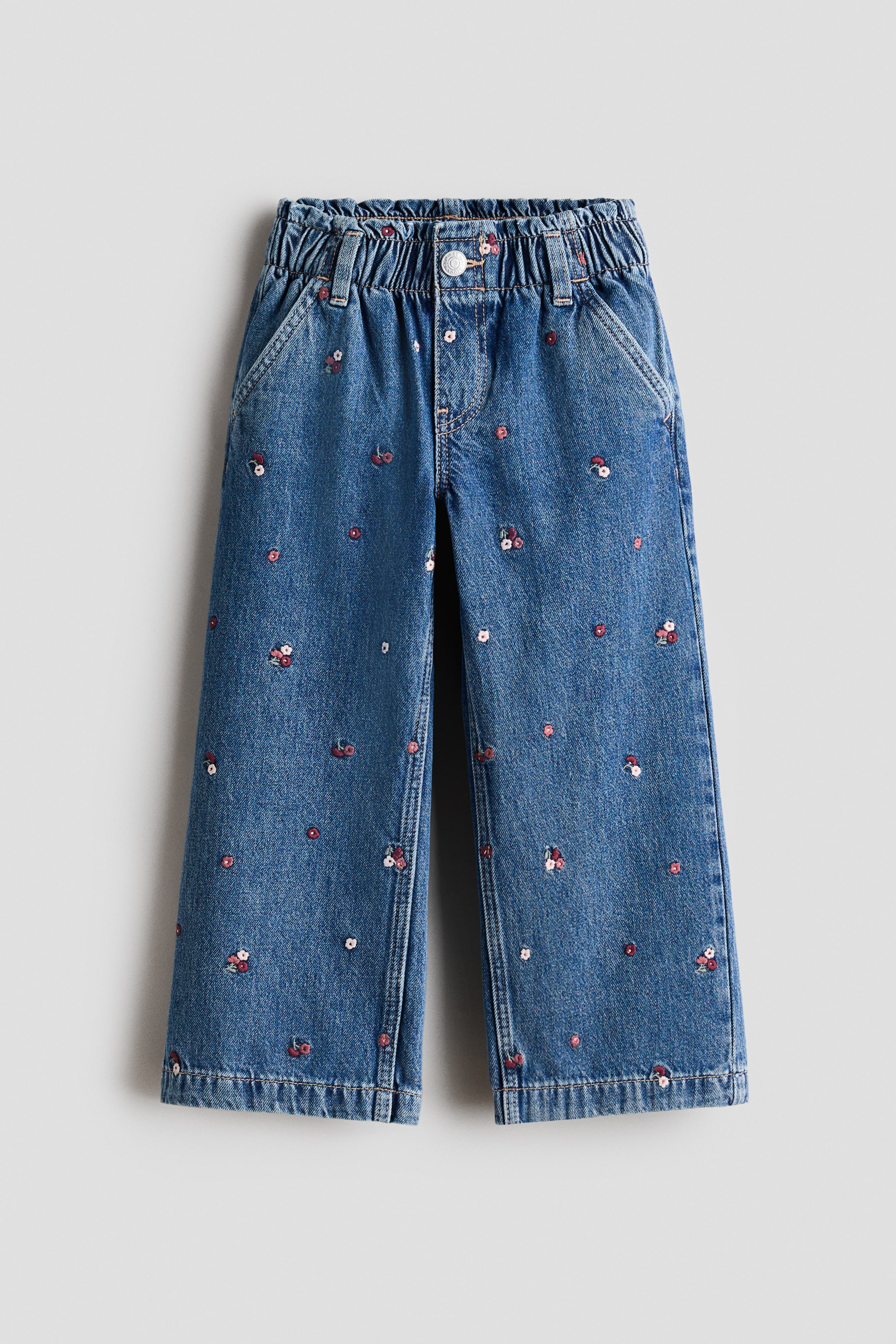 View larger image: These blue denim jeans are shown from the front, featuring a relaxed, wide-leg silhouette with a gathered paper-bag waist and a mock fly with a snap button. The denim features a scattered allover pattern of small embroidered flowers and cherries and includes diagonal side pockets.