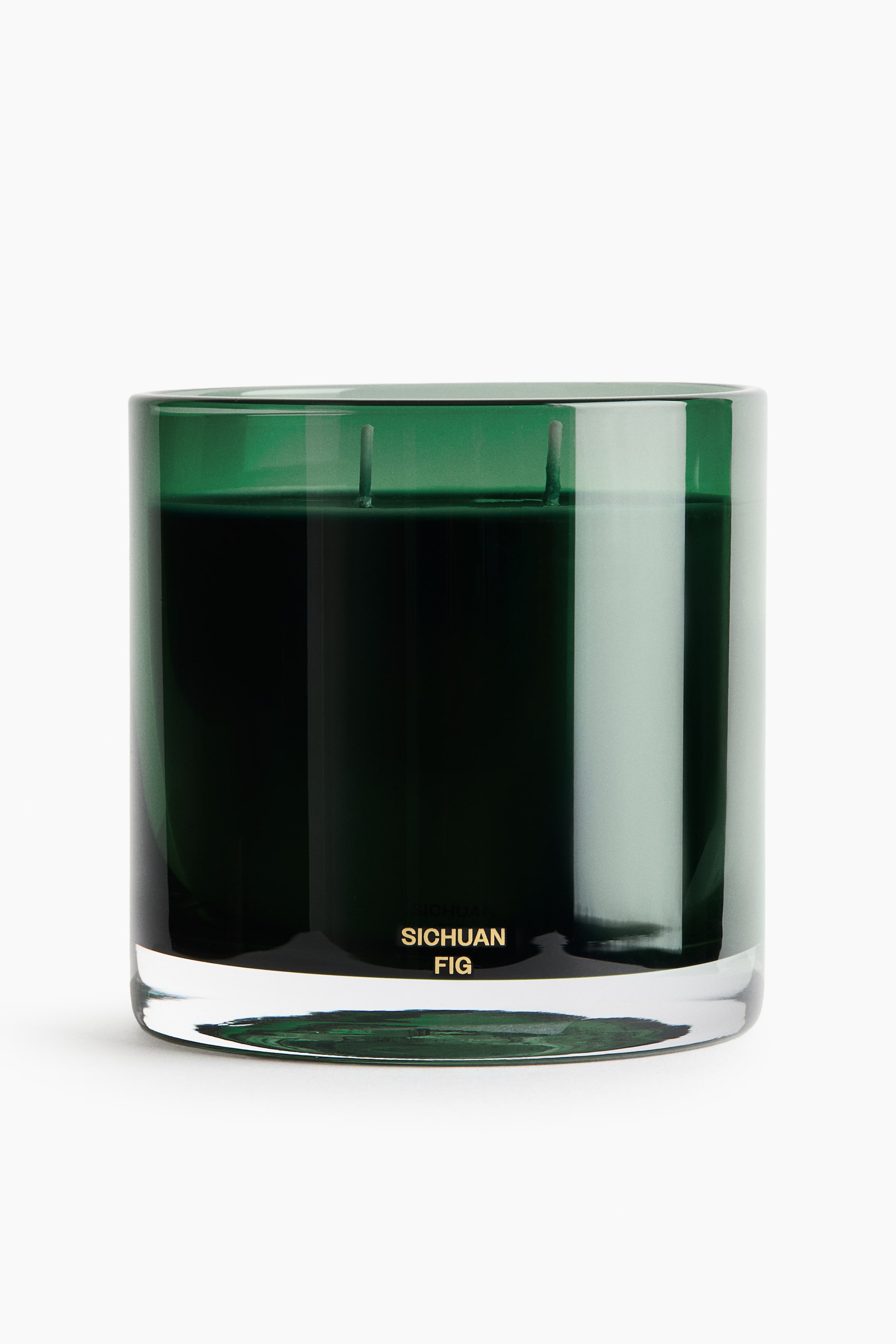 View larger image: A scented candle sits in a cylindrical dark green glass jar with a thick clear base, showing two wicks protruding from the dark wax. The front of the jar showcases the words "SICHUAN FIG" printed in gold lettering near the bottom.