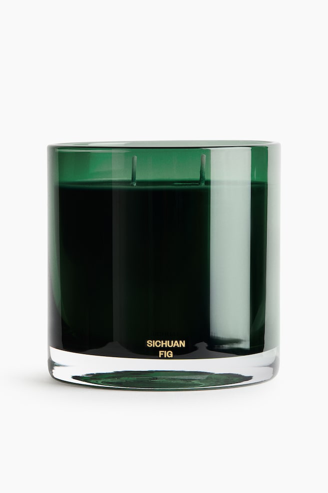Une bougie parfumée est placée dans un pot cylindrique en verre vert foncé avec une base épaisse et transparente, laissant apparaître deux mèches dépassant de la cire sombre. Lâavant du pot met en valeur les mots « SICHUAN FIG » imprimés en lettres dorées près du bas. - 1