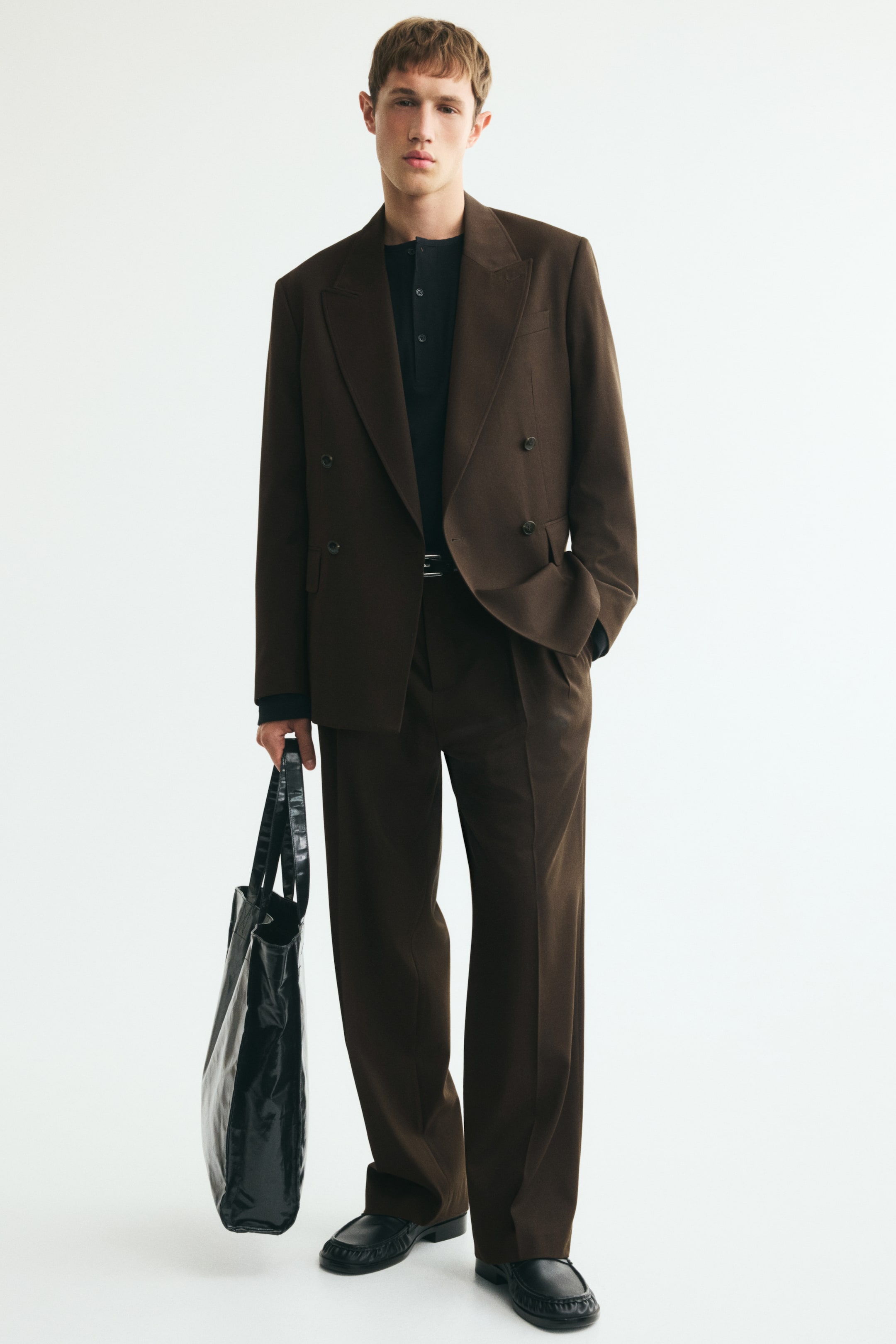 View larger image: Suit trousers in dark brown woven fabric featuring a loose fit and sharp front creases are being worn by the model. The trousers are styled with a matching double-breasted blazer, a tucked-in black polo shirt, black loafers, and a glossy black tote bag being held in one hand.