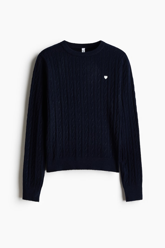 En marineblÃ¥ kabelstrikket sweater vises forfra med rund hals i ribstrik, lange Ã¦rmer med ribmanchetter og en ribkant forneden. Et lille broderet hvidt hjerte er placeret pÃ¥ venstre Ã¸verste bryst. - 1