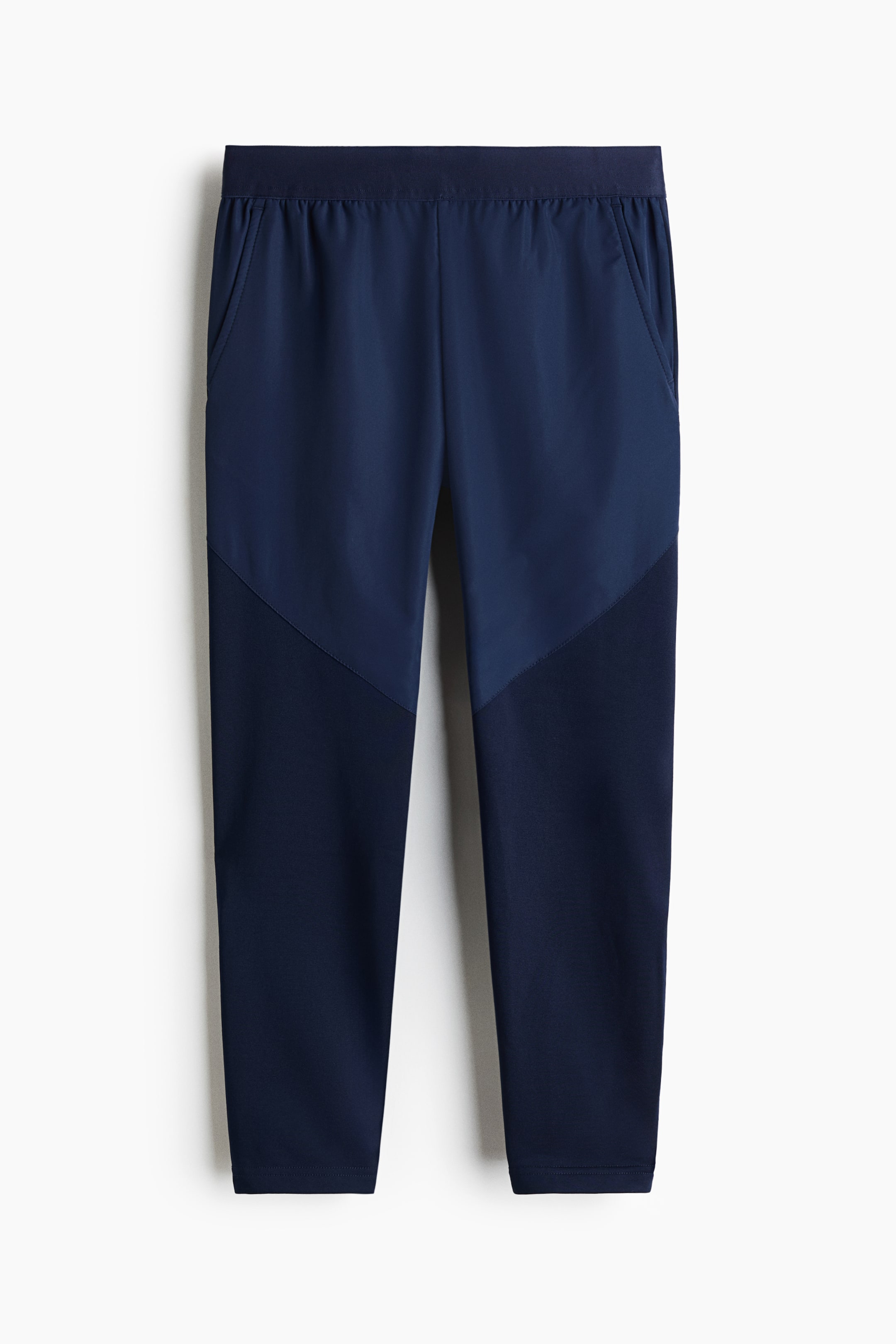 View larger image: These trousers boast a two-tone colour-block design in navy blue, with a lighter, smoother fabric on the upper section and a darker, subtly textured fabric on the lower section, separated by diagonal seams. An elasticated waistband and two side pockets are visible at the front.