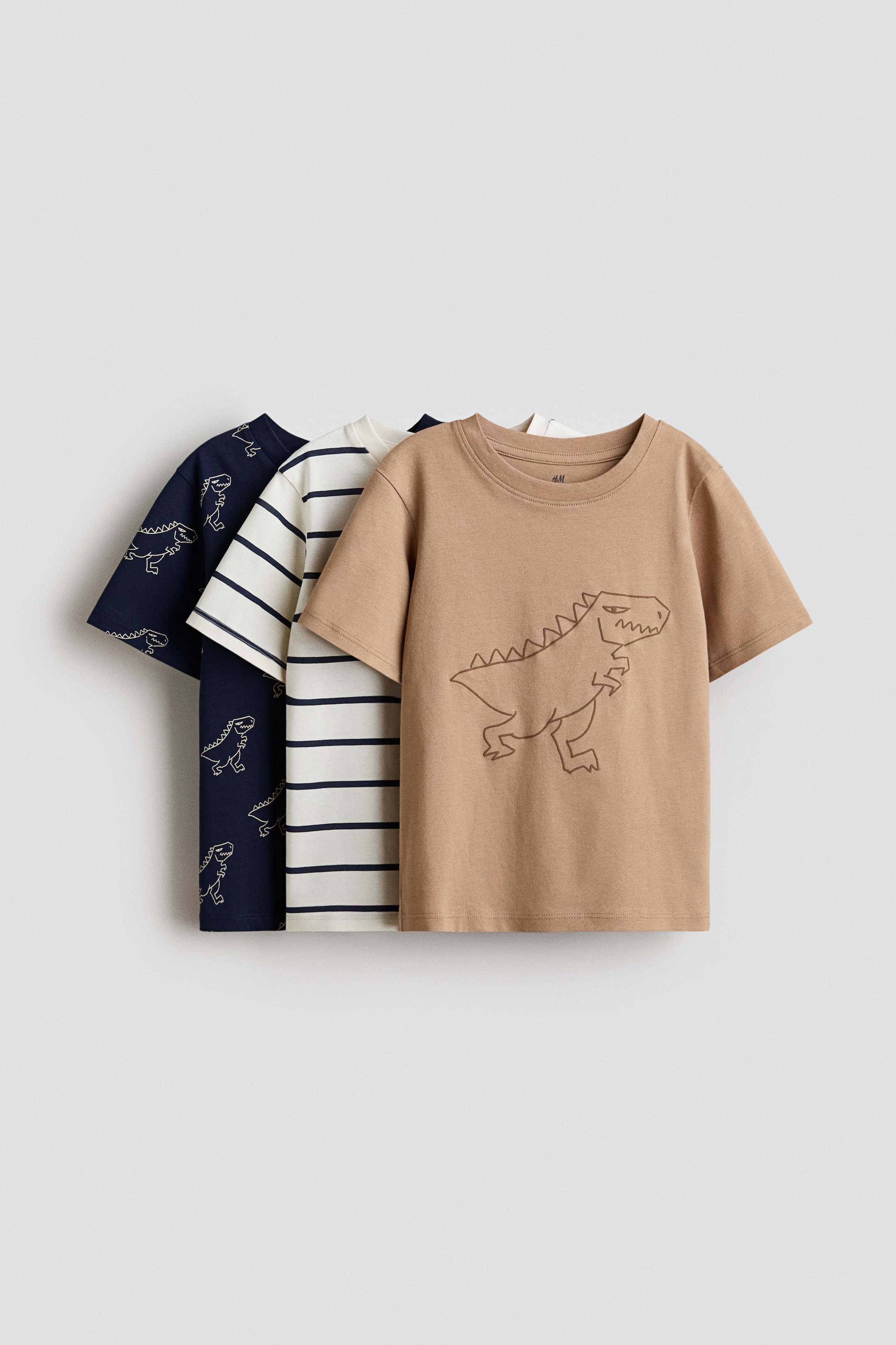 View larger image: Three classic t-shirts are being presented, layered with their fronts facing forward. The front t-shirt is tan with a brown dinosaur outline, the middle is off-white with navy stripes, and the back is navy with a white dinosaur pattern, all featuring round necklines and short sleeves.