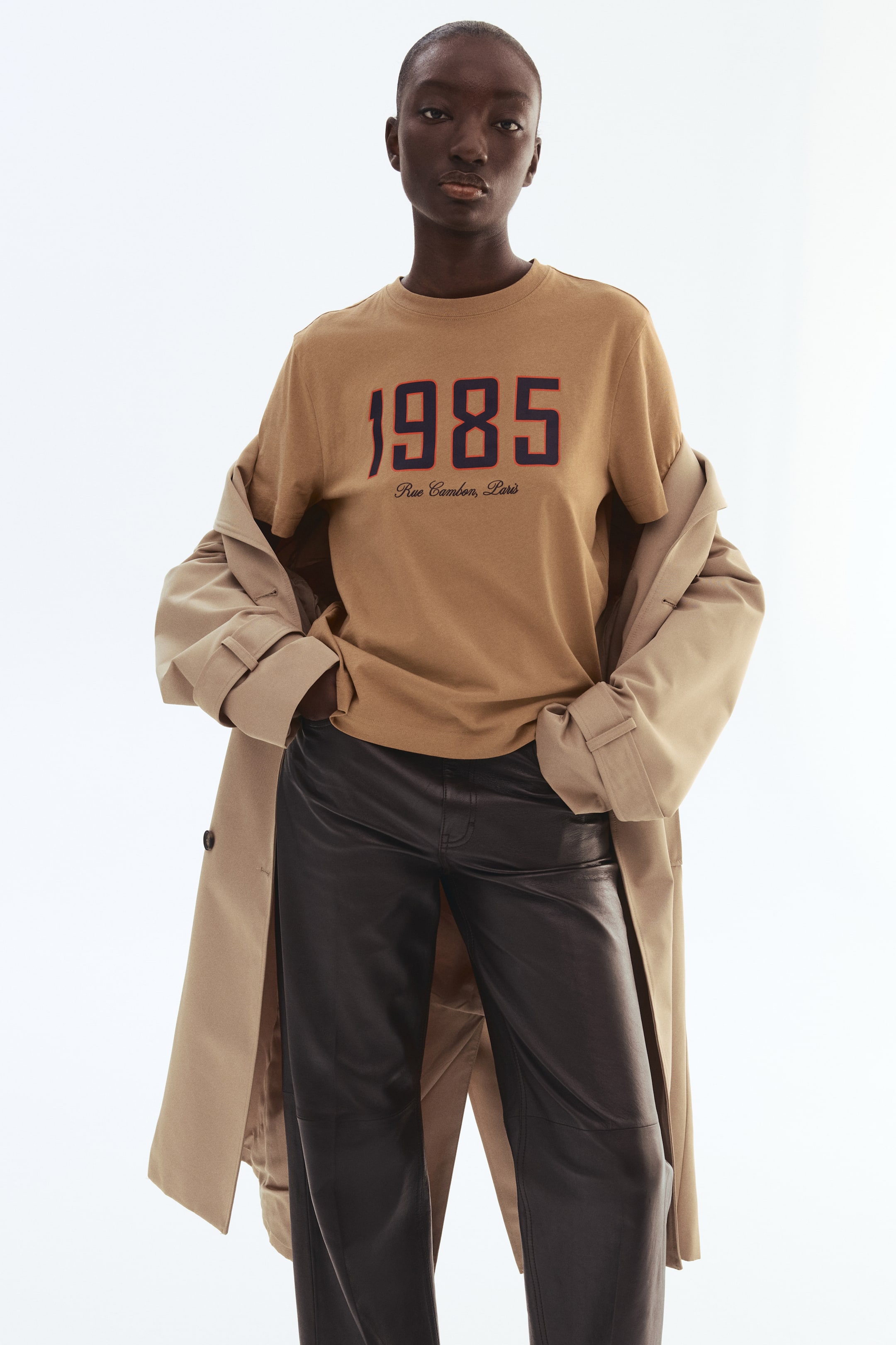 View larger image: A beige t-shirt showcasing "1985" in dark blue numerals with red outlining and "Rue Cambon, Paris" in script, styled alongside black leather trousers and a beige trench coat draped over the shoulders.