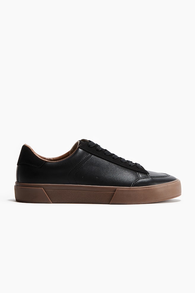 Une basket basse noire est présentée sous un angle latéral, avec une tige lisse en similicuir noir, un panneau de laçage mat noir et un contrefort de talon. Elle présente des lacets plats noirs, deux petites perforations sur le panneau latéral, et une semelle épaisse et plate de couleur marron avec un flanc texturé. - 1