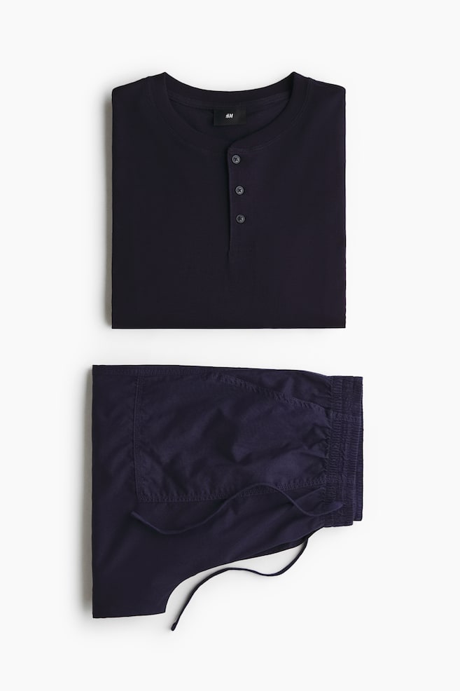 Ein gefaltetes dunkelmarineblaues Loungewear-Set zeigt eine Vorderansicht eines Henley-Shirts und der dazu passenden Hose. Das Oberteil verfÃ¼gt Ã¼ber einen Rundhalsausschnitt mit einer Dreiknopfleiste, und die gefaltete Hose zeigt einen elastischen Bund mit Kordelzug. - 1