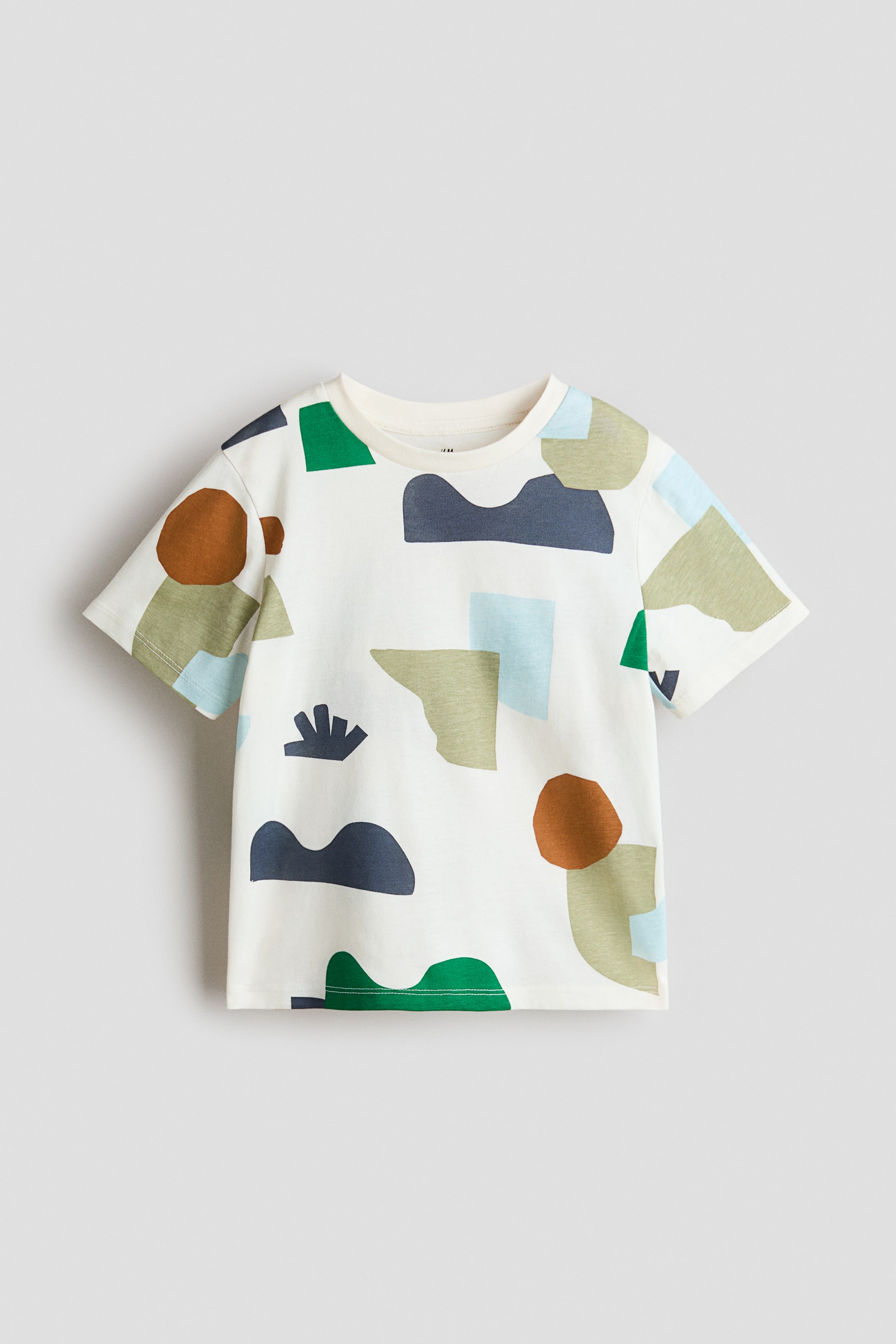 Agrandir l'image: Un t-shirt couleur crème est présenté de FACE, mettant en avant un motif abstrait de formes géométriques vertes, brunes, bleu clair et bleu foncé qui s’étend sur ses manches courtes et son col rond.