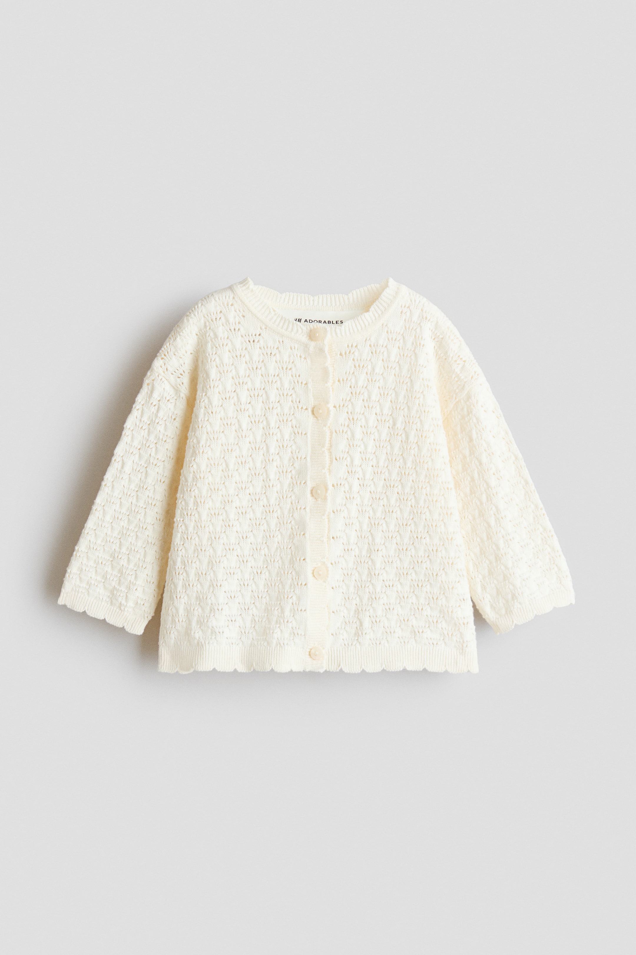 View larger image: FRONT: A cream cardigan features an open-knit pattern throughout, a round neckline, long sleeves, and a scalloped hem. Six light-colored buttons secure the front closure.
