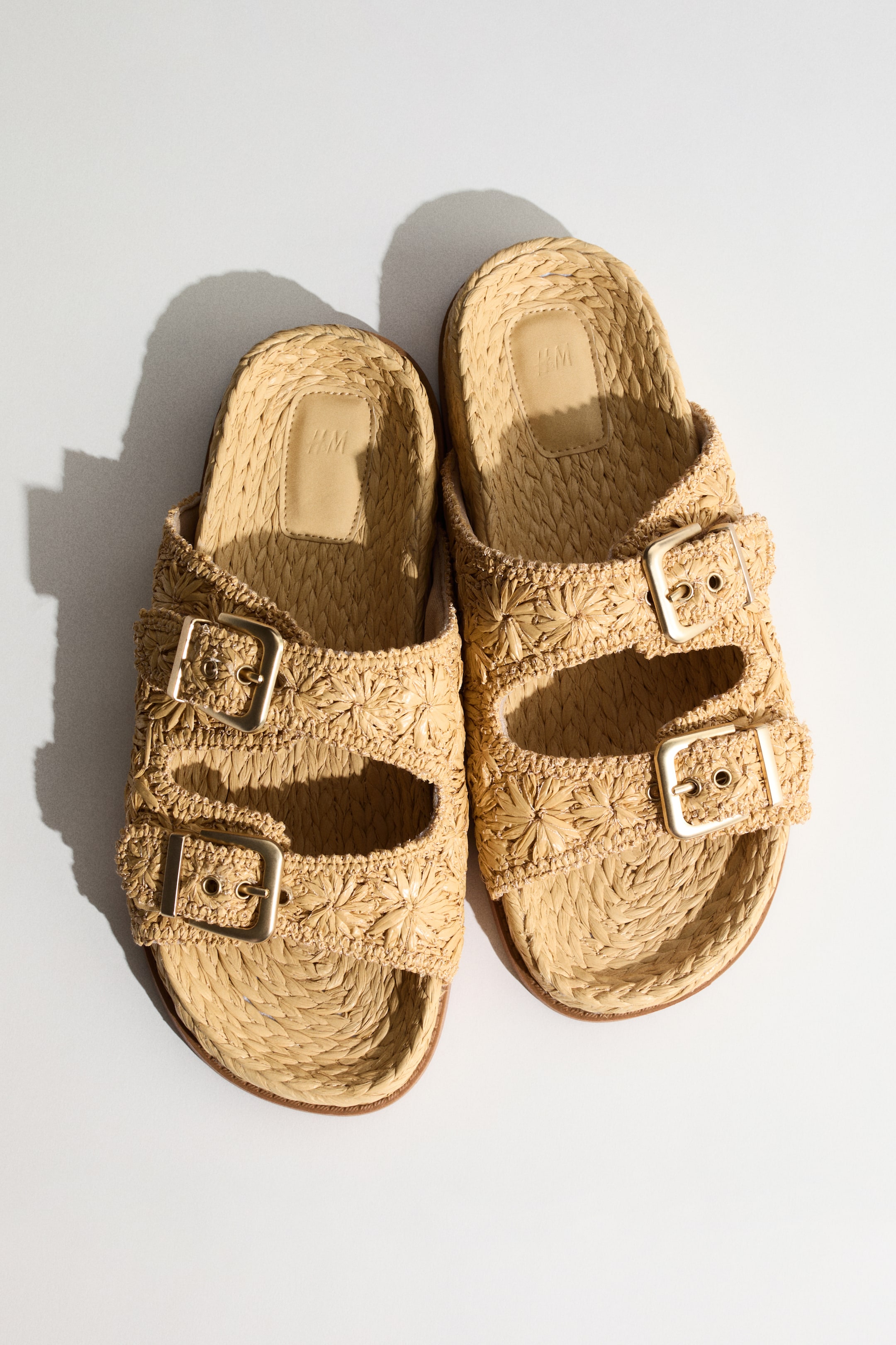 View larger image: A pair of light beige woven raffia slide sandals is positioned with the toes angled towards the upper left. These sandals feature two wide straps across the foot, embellished with gold-toned adjustable buckles and a woven starburst pattern, along with an intricately woven footbed.