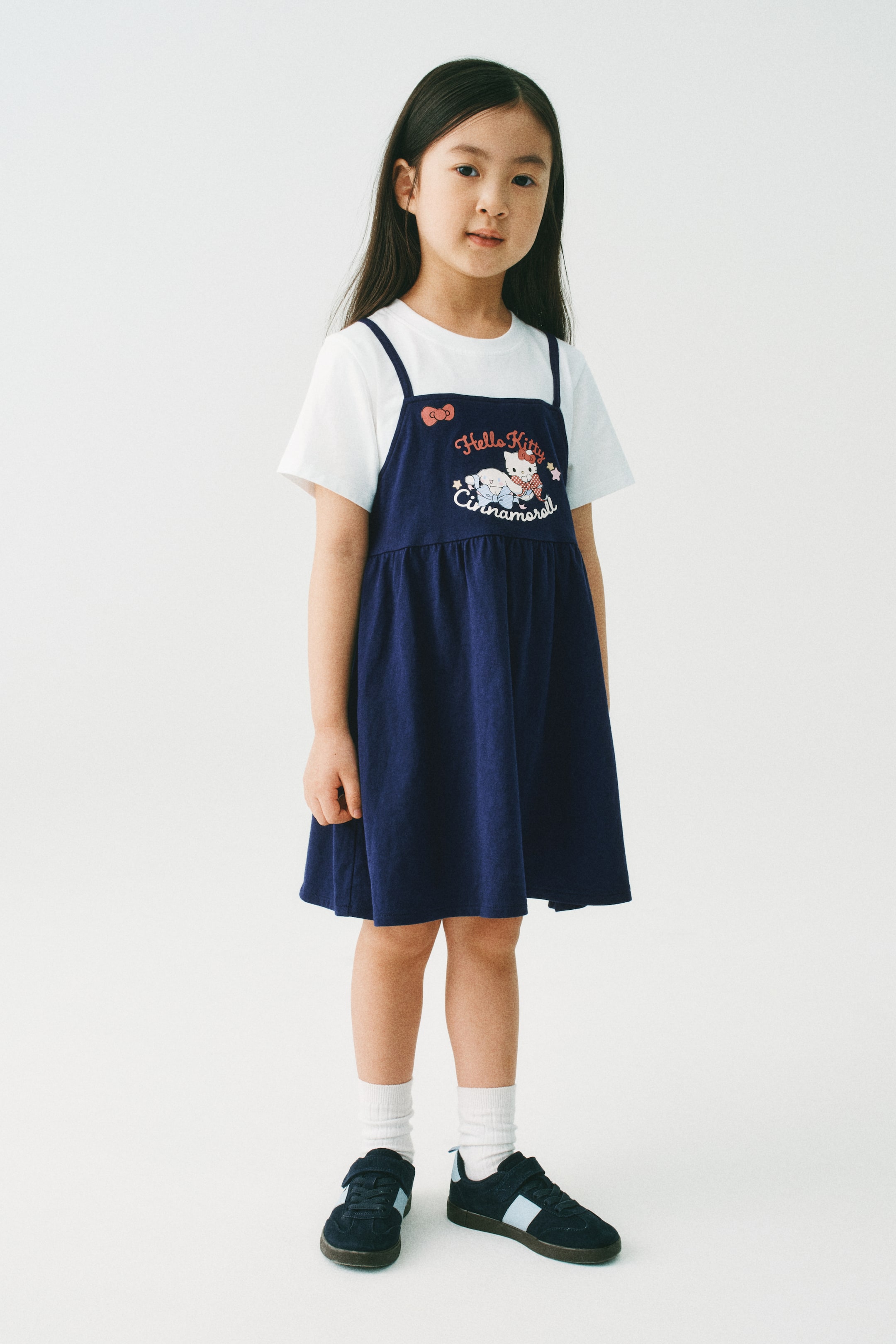 拡大画像を表示: モデルが着用しているのは、レイヤードドレスです。ネイビーのトップレイヤーは、スクエアネックライン、細いショルダーストラップ、ギャザーウエストシーム、そしてフロントプリントモチーフが特徴です。トップレイヤーの下には、ラウンドネックラインとリブトリムの短い袖が付いた白い縫い込みのアンダーレイヤーが見えています。