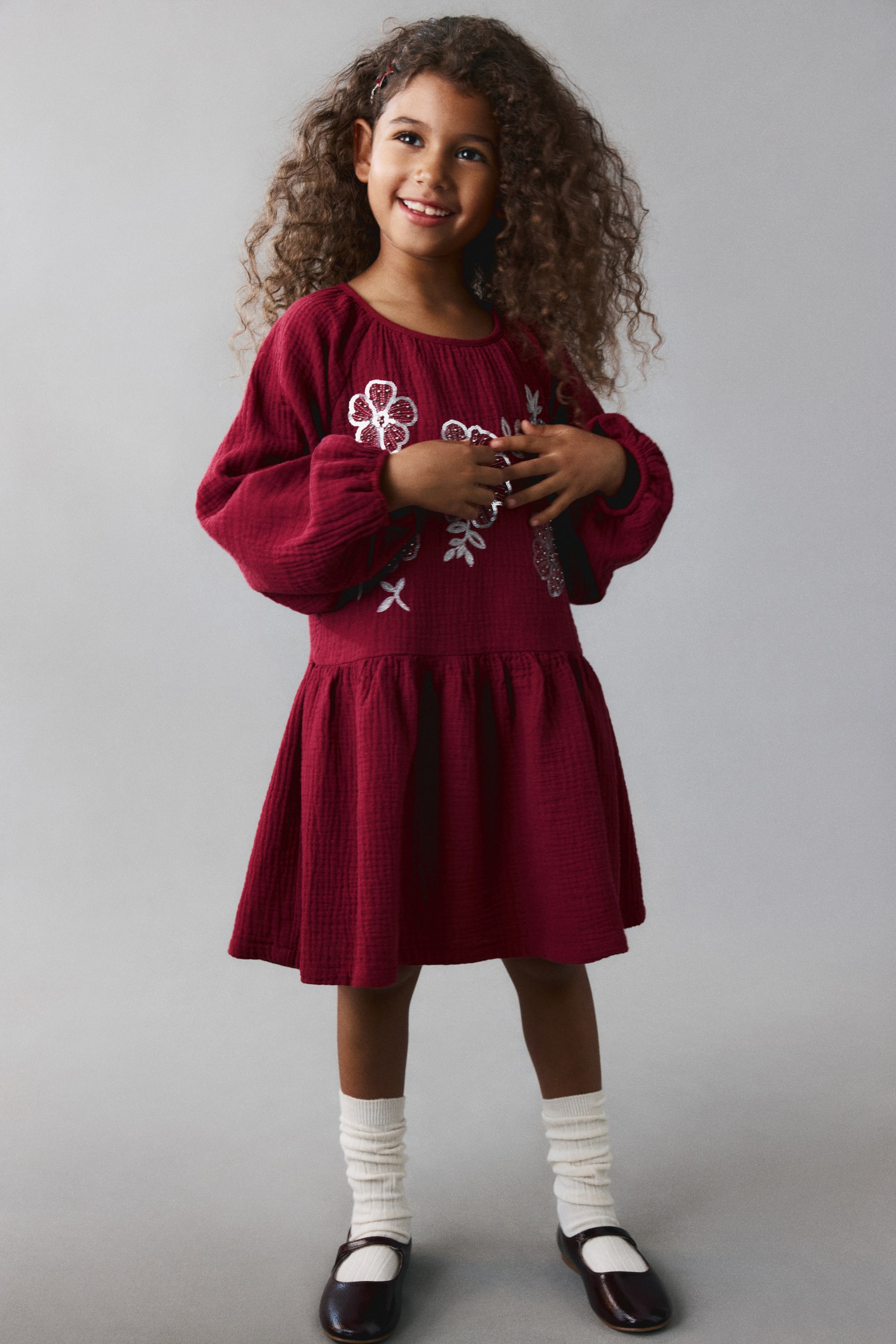 View larger image: A dark red crinkled cotton dress is shown from the front, featuring a round neckline, long raglan sleeves with gathered cuffs, and white floral sequin embroidery on the chest. The dress flows from a gathered waist seam into a flared skirt, styled with high white socks and dark Mary Jane shoes.